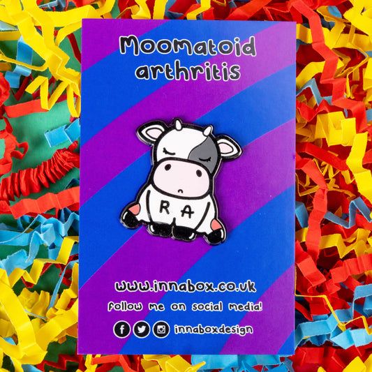 The Moomatoid Arthritis Cow Enamel Pin - Rheumatoid Arthritis on purple and blue stripe backing card on a red, blue and yellow coloured card confetti background. The sad cow shape pin has red marks on its lower back legs and the initials R and A across its middle. The hand drawn design is raising awareness for Rheumatoid Arthritis.