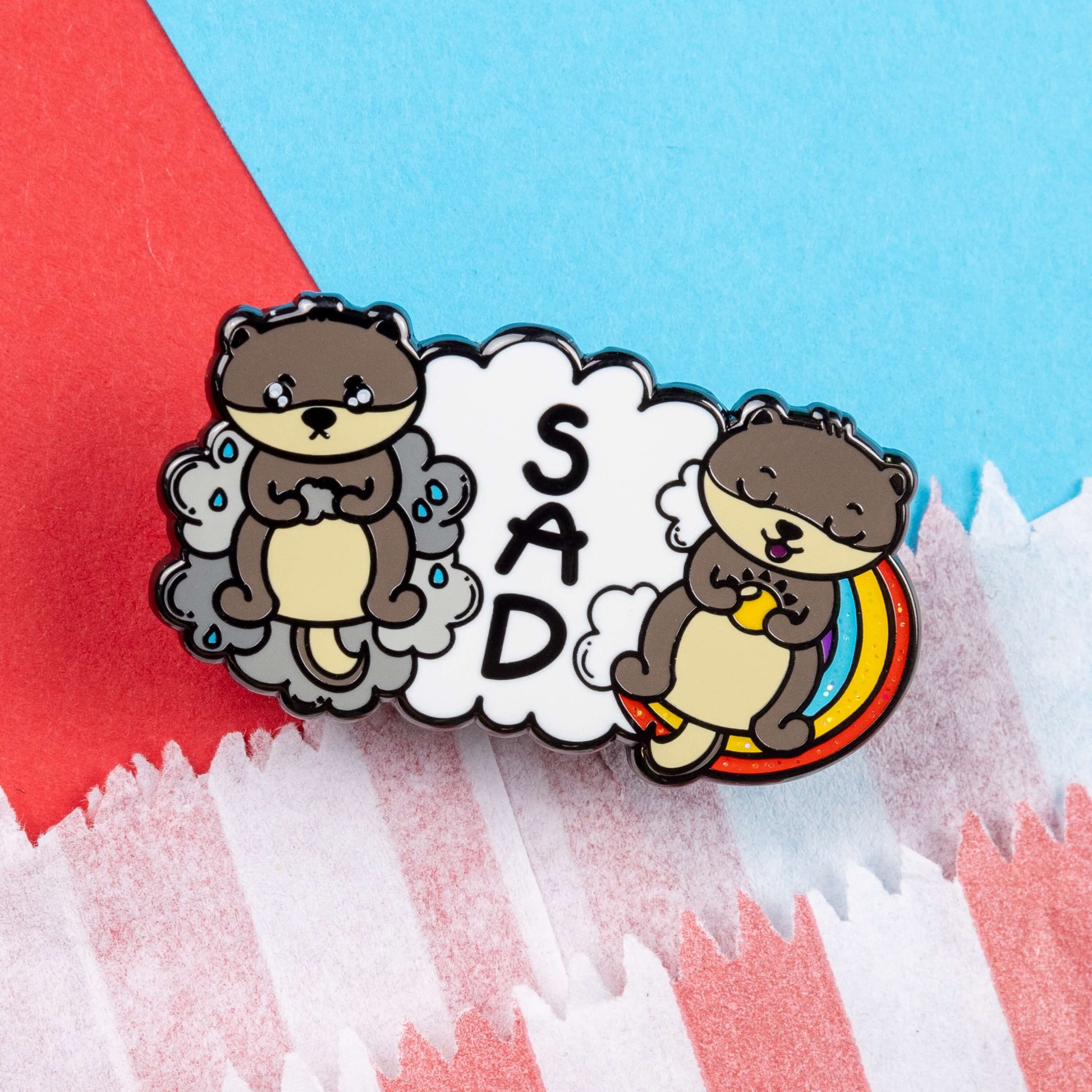The Seasonal Affective Otter Enamel Pin - Seasonal Affective Disorder SAD on a red and blue card background. The cloud shaped pin has two otters on either side, one sad on a raincloud clutching a raincloud and the other smiling on a rainbow clutching a sunshine. In the middle is the initials 'SAD'. The hand drawn design is raising awareness for Seasonal Affective Disorder.