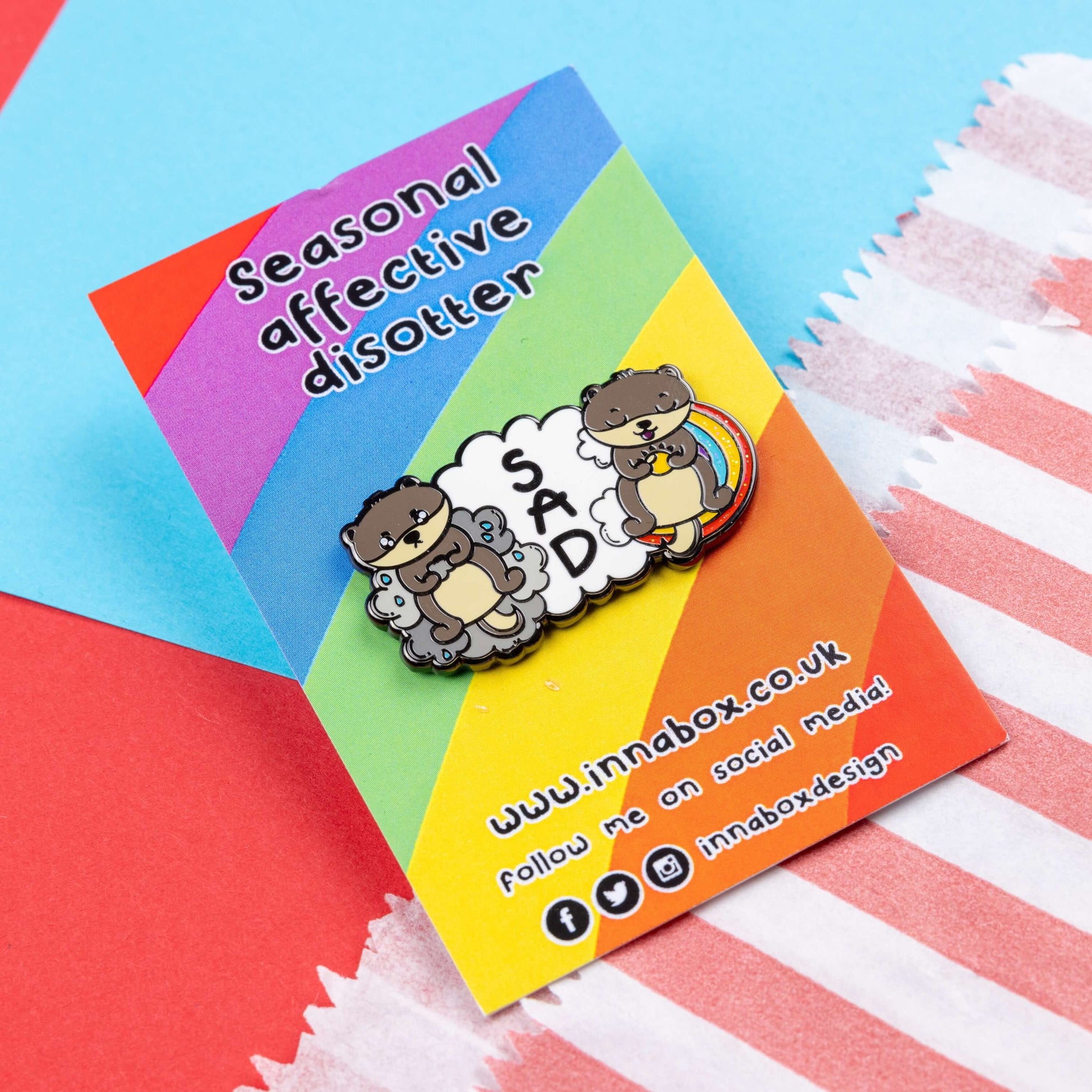 The Seasonal Affective Otter Enamel Pin - Seasonal Affective Disorder SAD on rainbow backing card laid on a red and blue card background. The cloud shaped pin has two otters on either side, one sad on a raincloud clutching a raincloud and the other smiling on a rainbow clutching a sunshine. In the middle is the initials 'SAD'. The hand drawn design is raising awareness for Seasonal Affective Disorder.