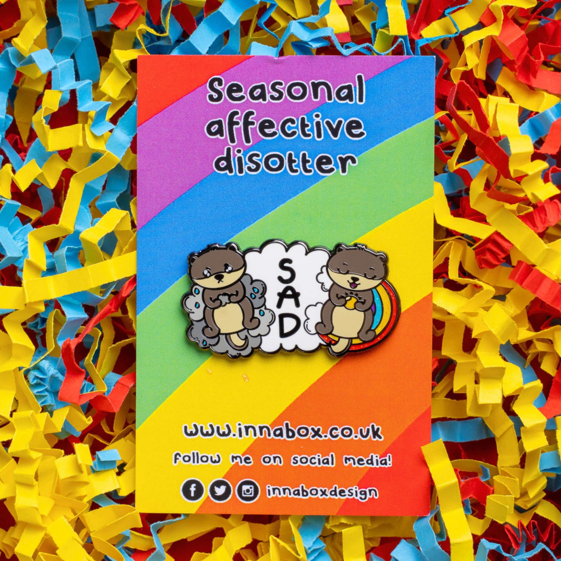 The Seasonal Affective Otter Enamel Pin - Seasonal Affective Disorder SAD on rainbow backing card laid on a red, yellow and blue card confetti background. The cloud shaped pin has two otters on either side, one sad on a raincloud clutching a raincloud and the other smiling on a rainbow clutching a sunshine. In the middle is the initials 'SAD'. The hand drawn design is raising awareness for Seasonal Affective Disorder.