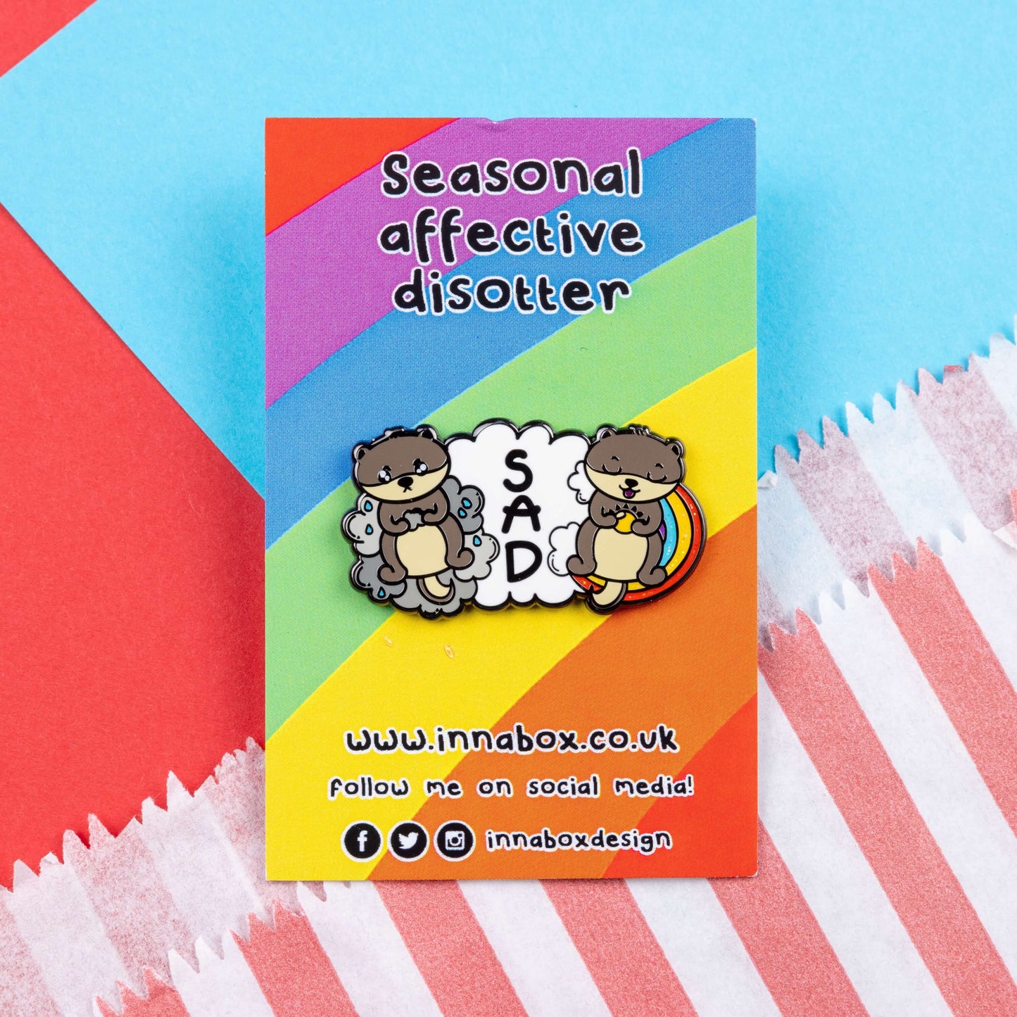 The Seasonal Affective Otter Enamel Pin - Seasonal Affective Disorder SAD on rainbow backing card laid on a red and blue card background. The cloud shaped pin has two otters on either side, one sad on a raincloud clutching a raincloud and the other smiling on a rainbow clutching a sunshine. In the middle is the initials 'SAD'. The hand drawn design is raising awareness for Seasonal Affective Disorder.