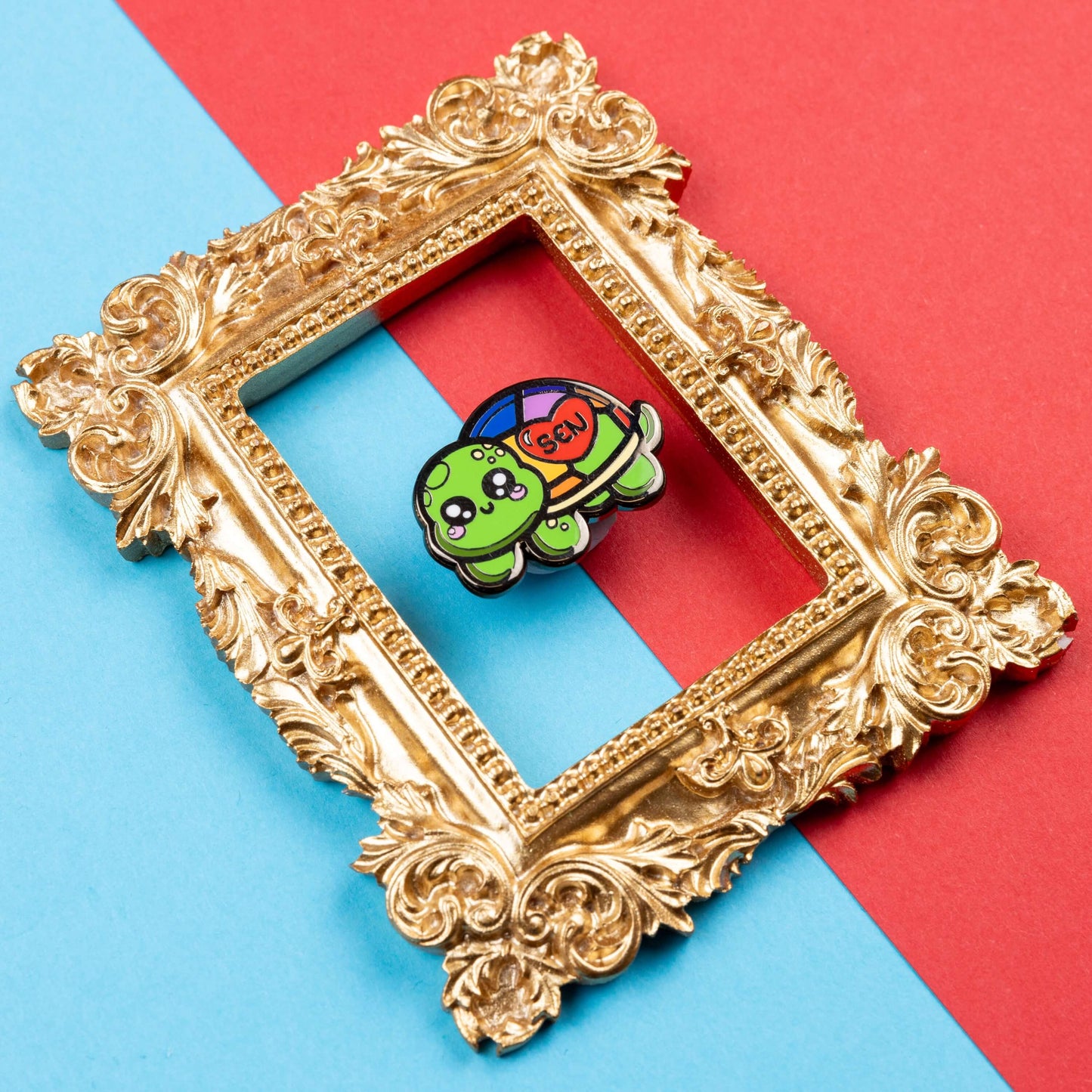 The Speshell Educational Needs Enamel Pin - SEN - Special Educational Needs on a red and blue background in a gold ornate frame. A rainbow shell kawaii cute style tortoise with pink cheeks and sparkling eyes, on its shell is a red heart with 'SEN' in the middle. The pin design is raising awareness for SEN Special Educational Needs.