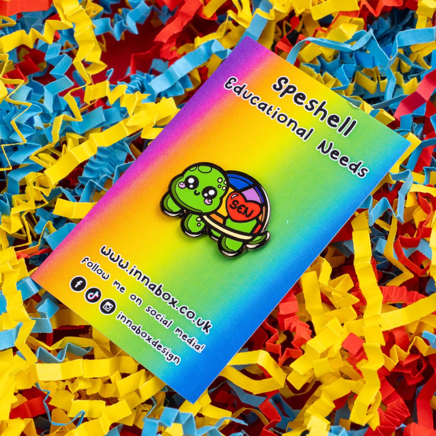 The Speshell Educational Needs Enamel Pin - SEN - Special Educational Needs on a rainbow backing card laid on a red, yellow and blue card confetti background. A rainbow shell kawaii cute style tortoise with pink cheeks and sparkling eyes, on its shell is a red heart with 'SEN' in the middle. The pin design is raising awareness for SEN Special Educational Needs.