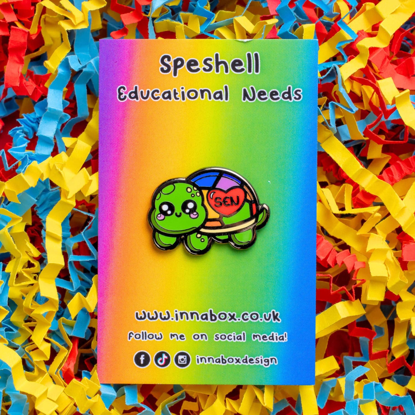 The Speshell Educational Needs Enamel Pin - SEN - Special Educational Needs on a rainbow backing card laid on a red, yellow and blue card confetti background. A rainbow shell kawaii cute style tortoise with pink cheeks and sparkling eyes, on its shell is a red heart with 'SEN' in the middle. The pin design is raising awareness for SEN Special Educational Needs.