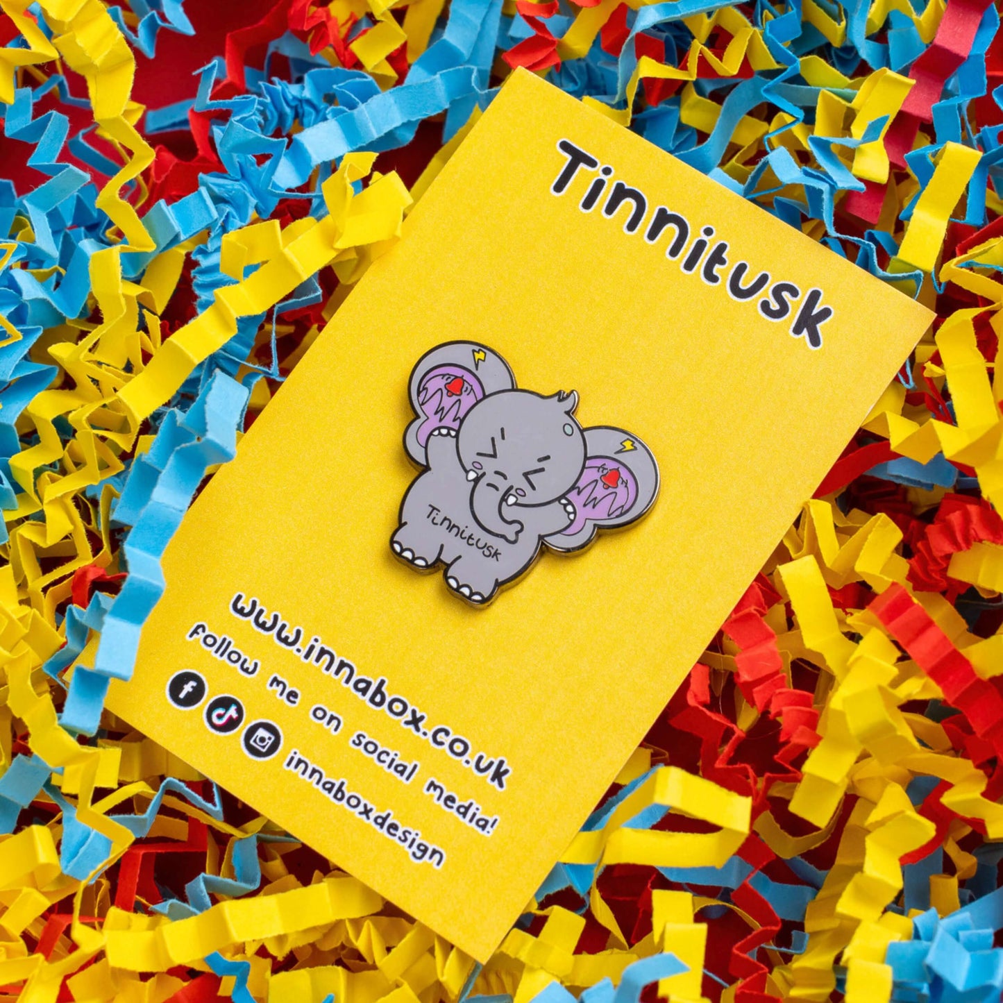 Tinnitus Enamel Pin on yellow backing card on a blue, yellow and red card confetti. A grey elephant shaped enamel pin with big ears with purple on the inside of them and black squiggly lines with red ringing alarm bells above the lines and yellow lightening bolts above the bells. The elephant has its eyes screwed shut and its arms up. 'Tinnitusk' is written in black across its middle.