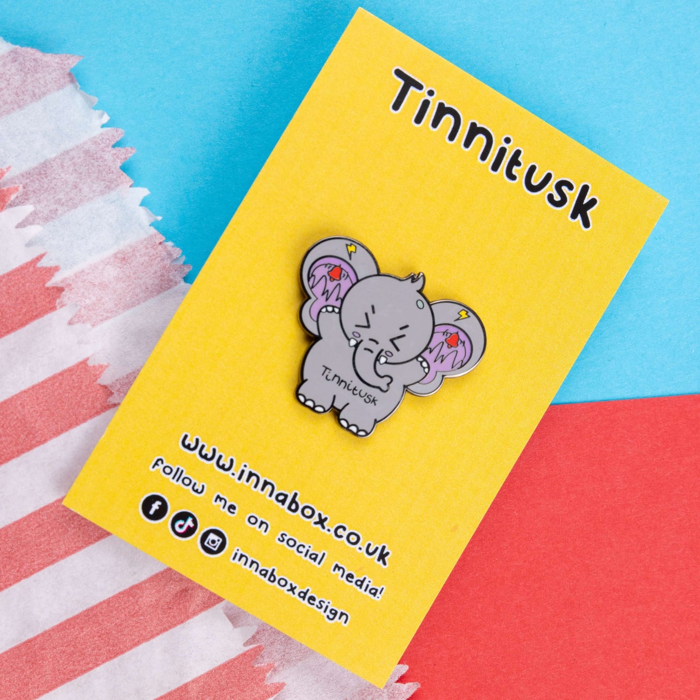 Tinnitus Enamel Pin on yellow backing card on a blue and red card. A grey elephant shaped enamel pin with big ears with purple on the inside of them and black squiggly lines with red ringing alarm bells above the lines and yellow lightening bolts above the bells. The elephant has its eyes screwed shut and its arms up. 'Tinnitusk' is written in black across its middle.