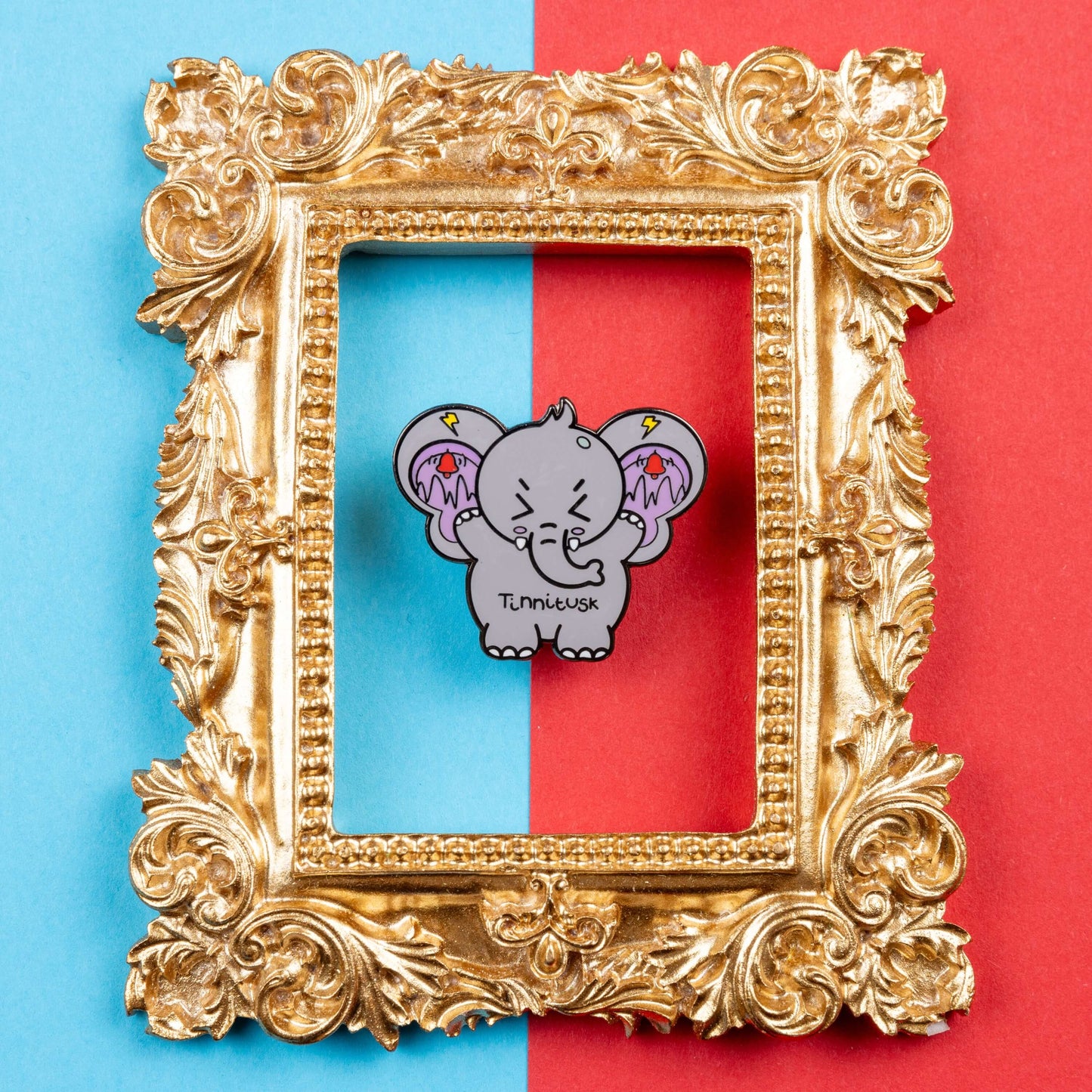 Tinnitus Enamel Pin on blue and red card inside a gold ornate frame. A grey elephant shaped enamel pin with big ears with purple on the inside of them and black squiggly lines with red ringing alarm bells above the lines and yellow lightening bolts above the bells. The elephant has its eyes screwed shut and its arms up. 'Tinnitusk' is written in black across its middle.