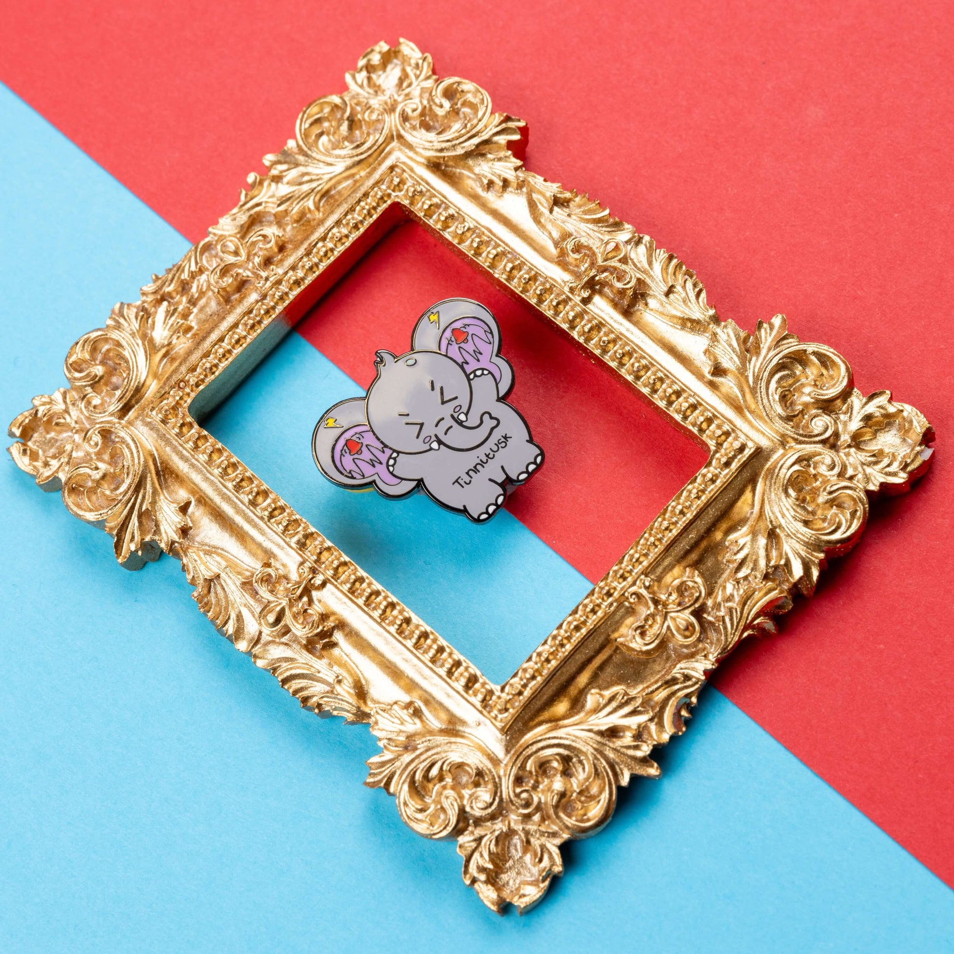 Tinnitus Enamel Pin on blue and red card inside a gold ornate frame. A grey elephant shaped enamel pin with big ears with purple on the inside of them and black squiggly lines with red ringing alarm bells above the lines and yellow lightening bolts above the bells. The elephant has its eyes screwed shut and its arms up. 'Tinnitusk' is written in black across its middle.
