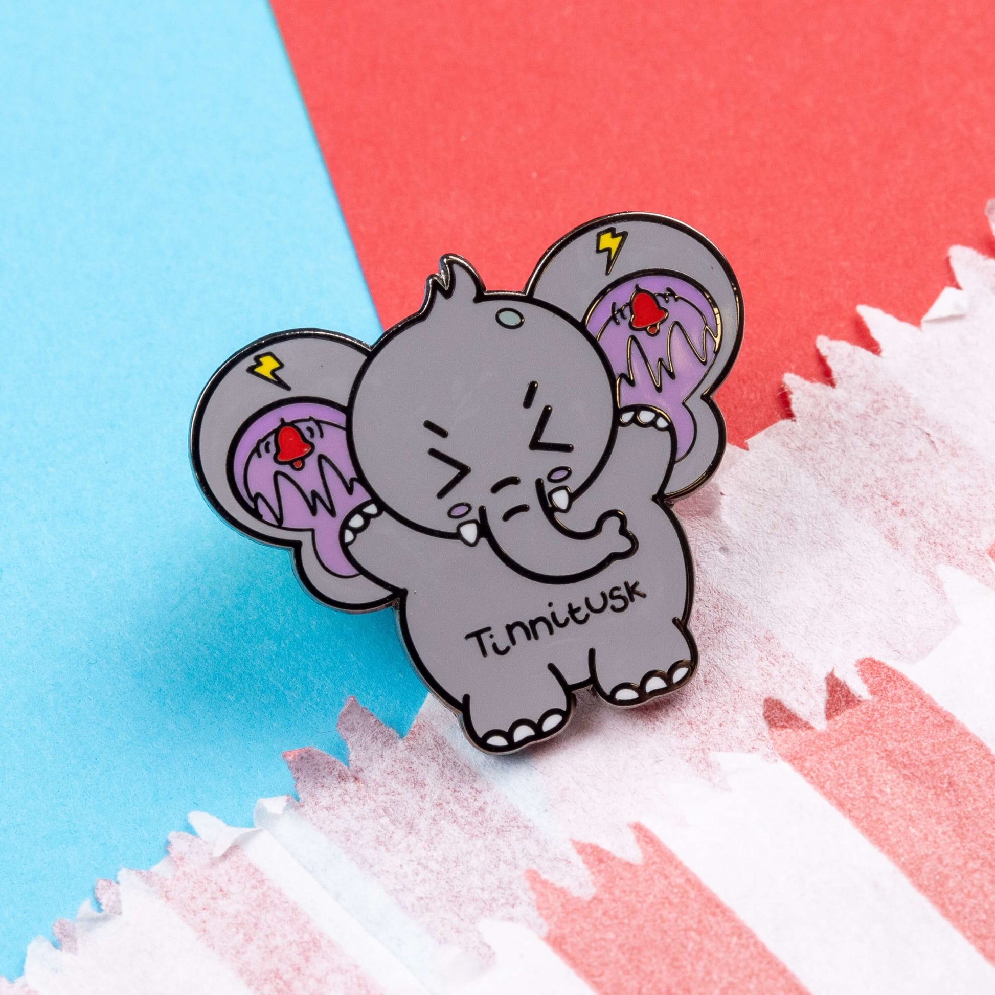 Tinnitus Enamel Pin on blue and red card. A grey elephant shaped enamel pin with big ears with purple on the inside of them and black squiggly lines with red ringing alarm bells above the lines and yellow lightening bolts above the bells. The elephant has its eyes screwed shut and its arms up. 'Tinnitusk' is written in black across its middle.