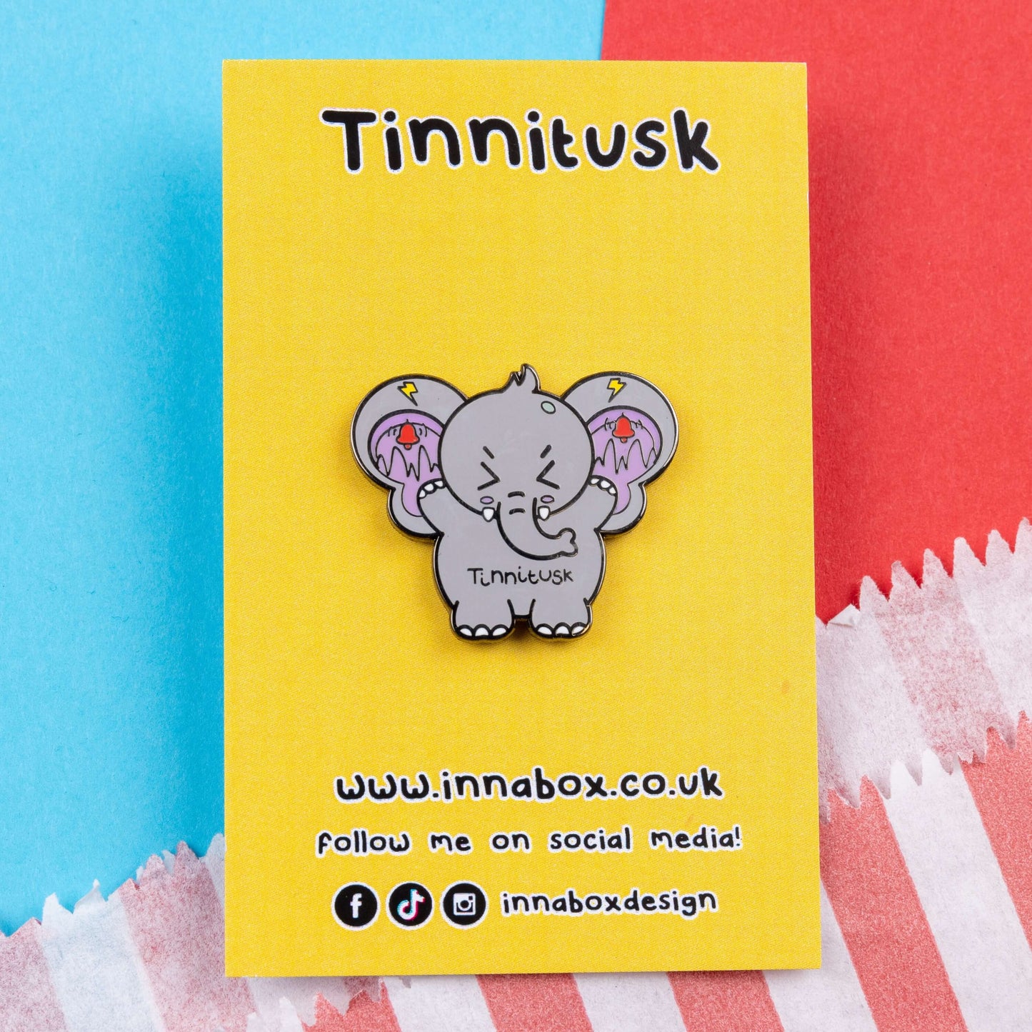 Tinnitus Enamel Pin on yellow backing card on a blue and red card. A grey elephant shaped enamel pin with big ears with purple on the inside of them and black squiggly lines with red ringing alarm bells above the lines and yellow lightening bolts above the bells. The elephant has its eyes screwed shut and its arms up. 'Tinnitusk' is written in black across its middle.