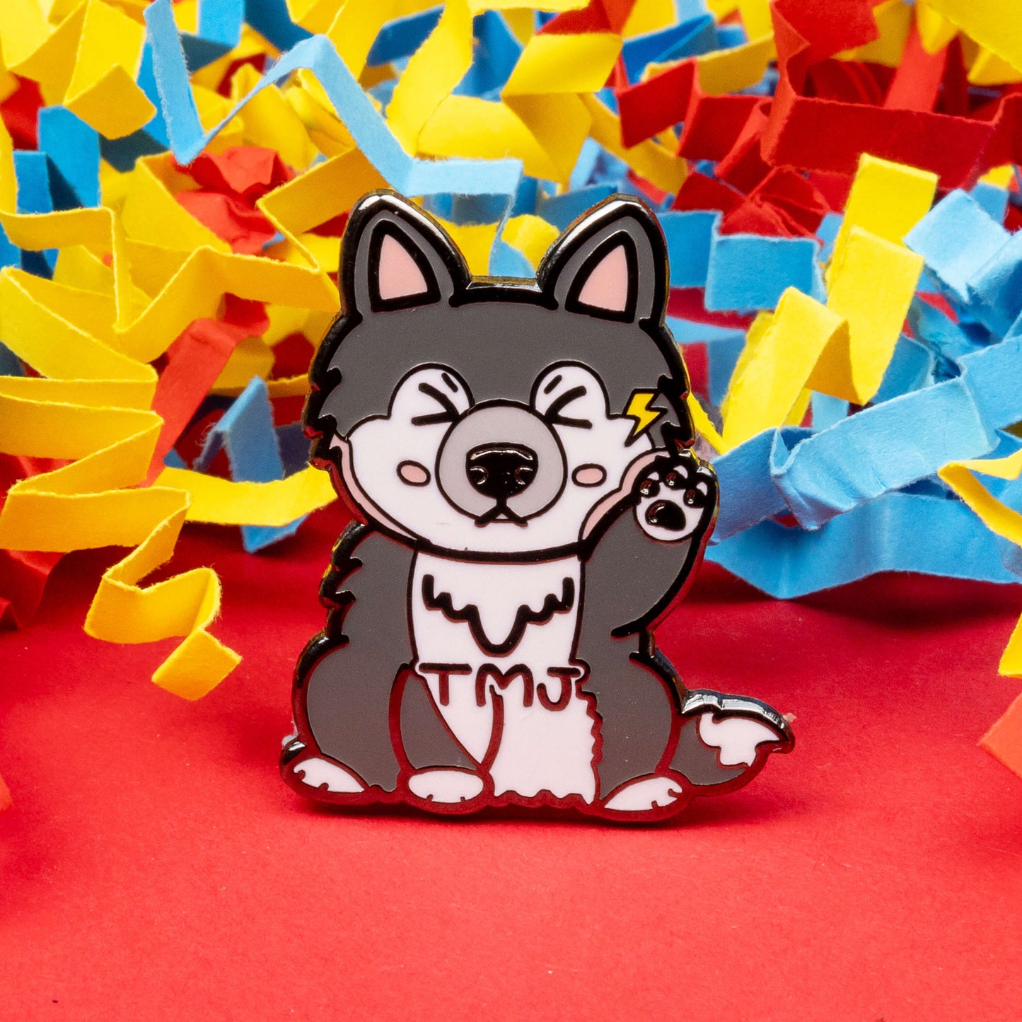 The Tempawromandibular Disorder Dog Enamel Pin - Temporomandibular Disorder TMJ on a red background with coloured card confetti. The grey husky dog shaped enamel pin has a pained face with shut eyes, one paw up by its jaw with a yellow lightning bolt and black text across its chest reading 'TMJ'. The hand drawn design is raising awareness for Temporomandibular Disorder TMJ.