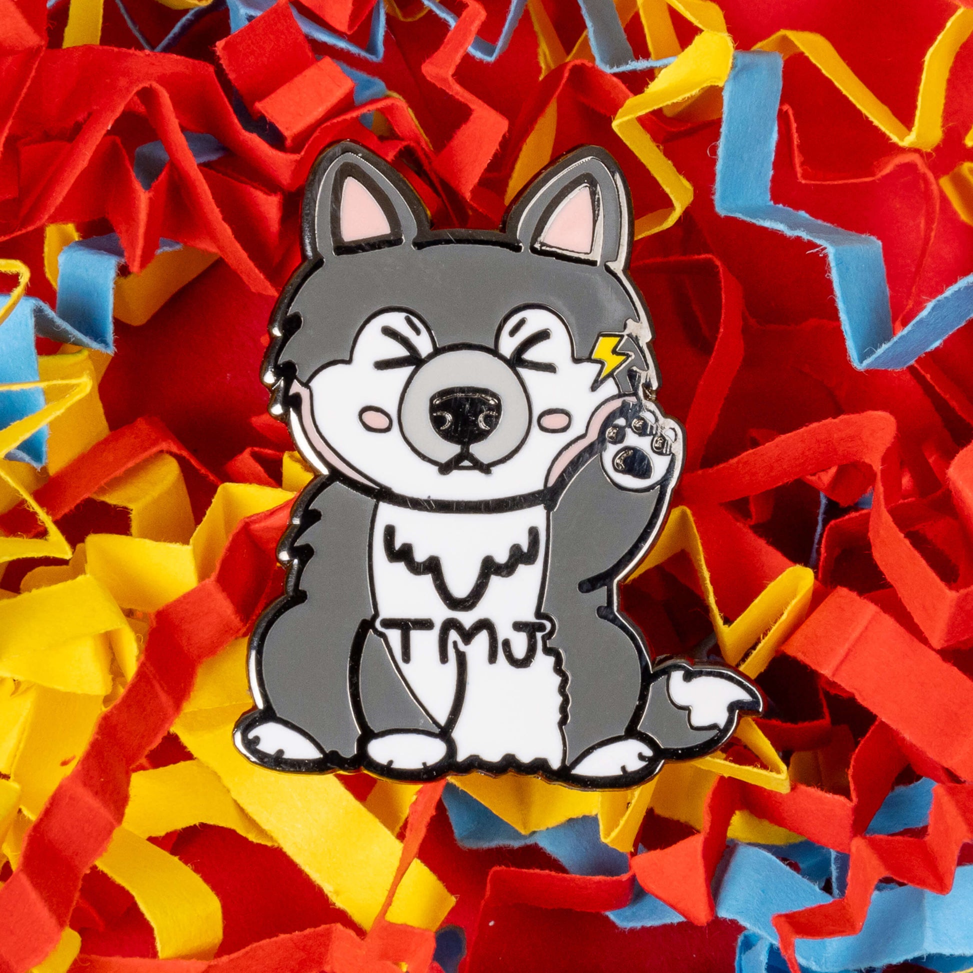 The Tempawromandibular Disorder Dog Enamel Pin - Temporomandibular Disorder TMJ on a red, blue and yellow coloured card confetti background. The grey husky dog shaped enamel pin has a pained face with shut eyes, one paw up by its jaw with a yellow lightning bolt and black text across its chest reading 'TMJ'. The hand drawn design is raising awareness for Temporomandibular Disorder TMJ.