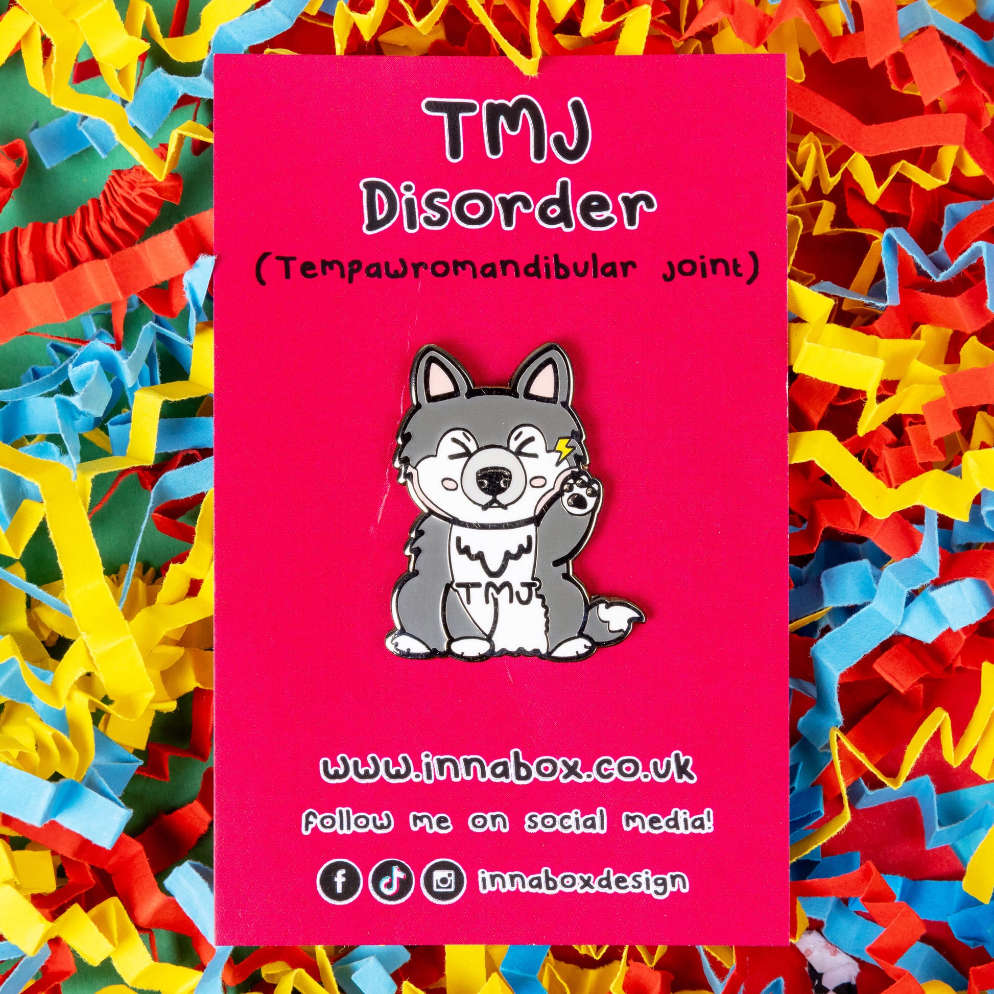 The Tempawromandibular Disorder Dog Enamel Pin - Temporomandibular Disorder TMJ on red backing card on blue, yellow and red card confetti background. The grey husky dog shaped enamel pin has a pained face with shut eyes, one paw up by its jaw with a yellow lightning bolt and black text across its chest reading 'TMJ'. The hand drawn design is raising awareness for Temporomandibular Disorder TMJ.