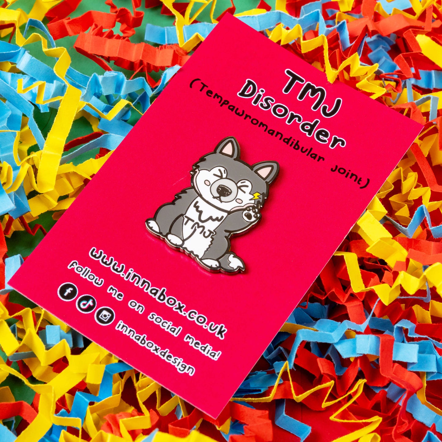 The Tempawromandibular Disorder Dog Enamel Pin - Temporomandibular Disorder TMJ on red backing card on blue, yellow and red card confetti background. The grey husky dog shaped enamel pin has a pained face with shut eyes, one paw up by its jaw with a yellow lightning bolt and black text across its chest reading 'TMJ'. The hand drawn design is raising awareness for Temporomandibular Disorder TMJ.