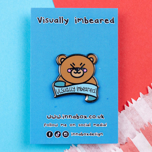 The Visually Imbeared Bear Enamel Pin - Visually Impaired on blue backing card laid on a red and blue card background. The brown teddy bear shaped pin has its eyes scrunched shut smiling with pink cheeks, underneath is a blue banner with black text reading 'visually imbeared'. The hand drawn design is raising awareness for visual impairment and blindness.