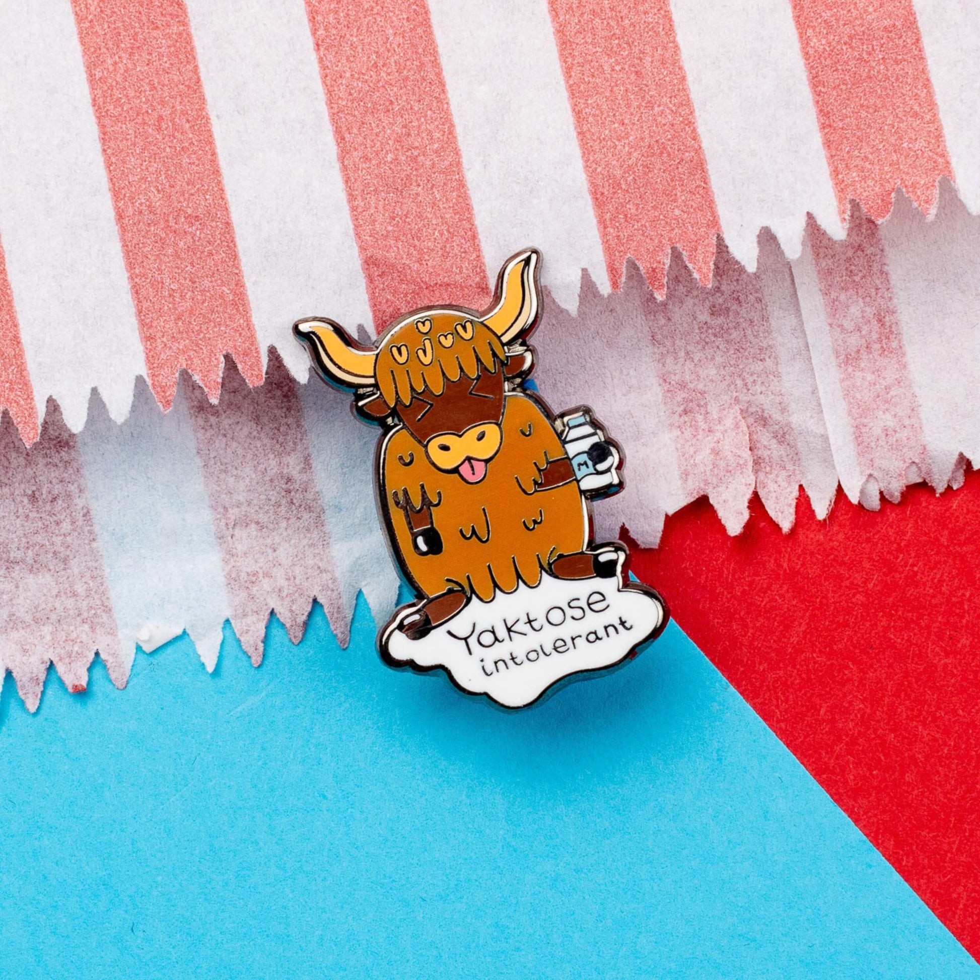 Yaktose Intolerant Enamel Pin - Lactose Intolerant on a blue and red background. The enamel pin is a yak sat holding a bottle of milk with it's tongue out looking disgusted. There is a puddle of milk under the yak with black text that reads 'yaktose intolerant'. The hand drawn design is made to raise awareness for lactose intolerance.