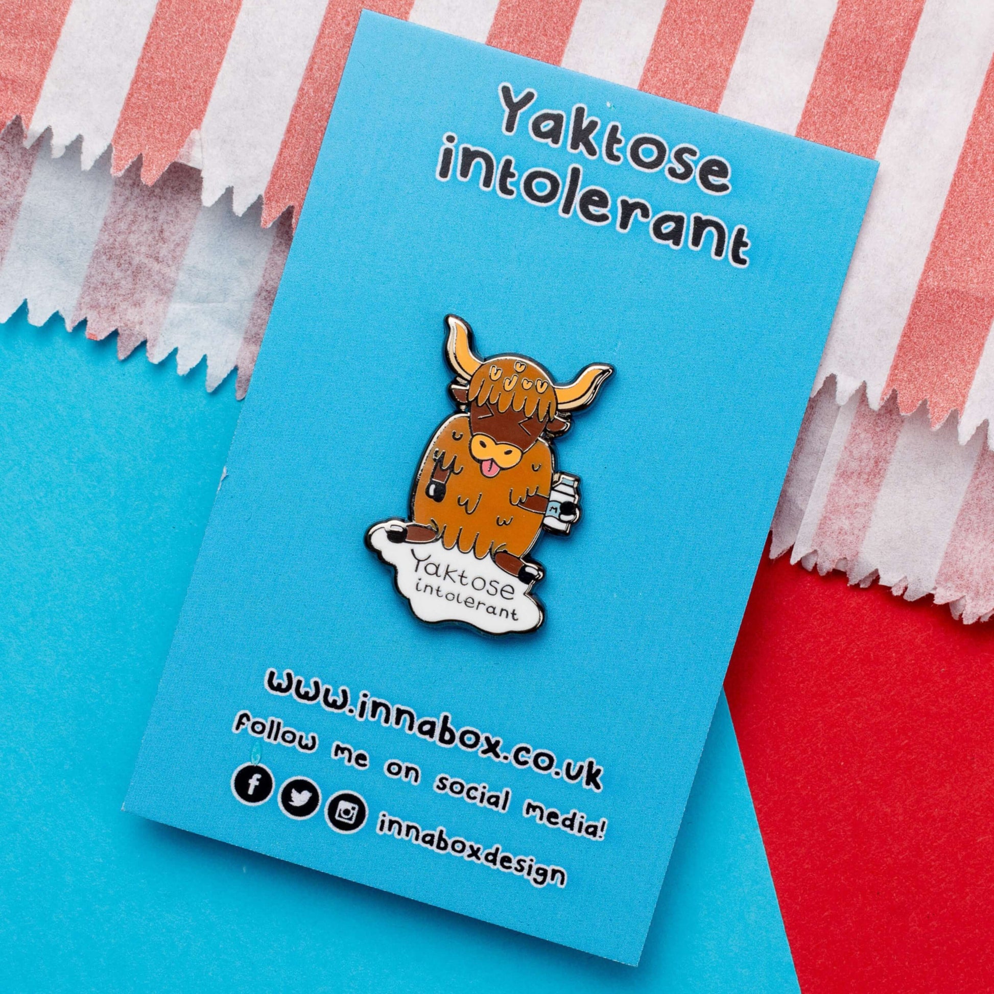 Yaktose Intolerant Enamel Pin - Lactose Intolerant on blue backing card laying on a blue and red background. The enamel pin is a yak sat holding a bottle of milk with it's tongue out looking disgusted. There is a puddle of milk under the yak with black text that reads 'yaktose intolerant'. The hand drawn design is made to raise awareness for lactose intolerance.