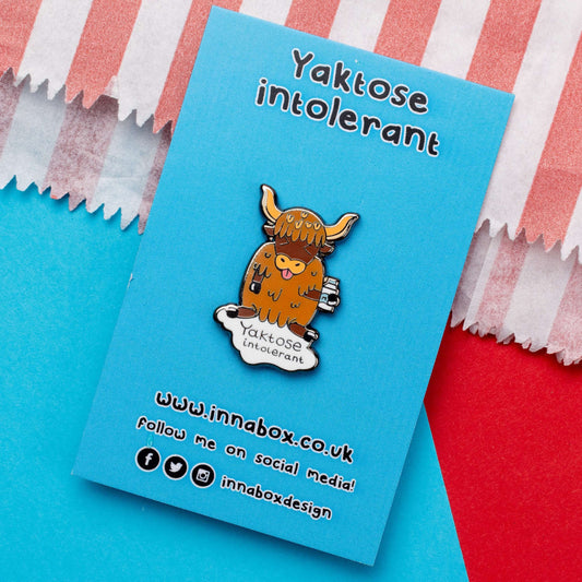 Yaktose Intolerant Enamel Pin - Lactose Intolerant on blue backing card laying on a blue and red background. The enamel pin is a yak sat holding a bottle of milk with it's tongue out looking disgusted. There is a puddle of milk under the yak with black text that reads 'yaktose intolerant'. The hand drawn design is made to raise awareness for lactose intolerance.