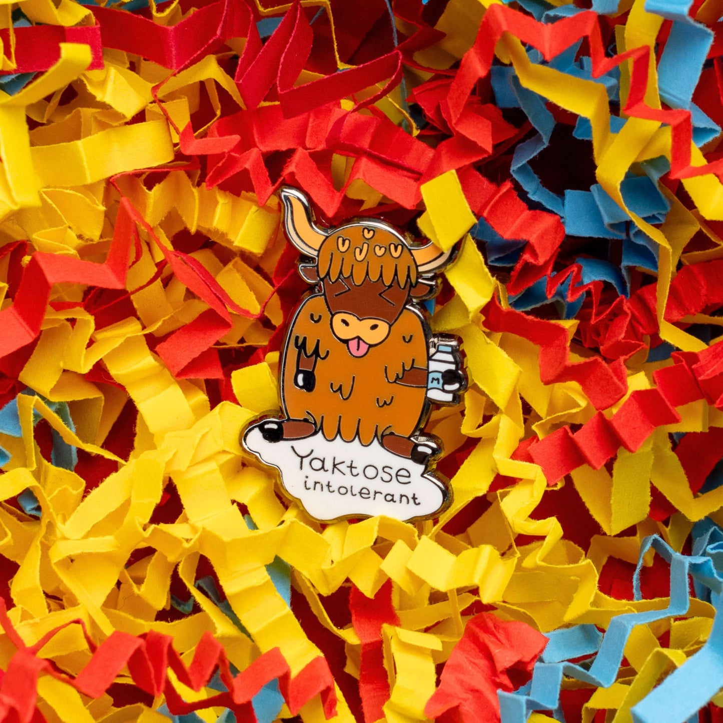 Yaktose Intolerant Enamel Pin - Lactose Intolerant on a blue, red and yellow crinkle card confetti background. The enamel pin is a yak sat holding a bottle of milk with it's tongue out looking disgusted. There is a puddle of milk under the yak with black text that reads 'yaktose intolerant'. The hand drawn design is made to raise awareness for lactose intolerance.