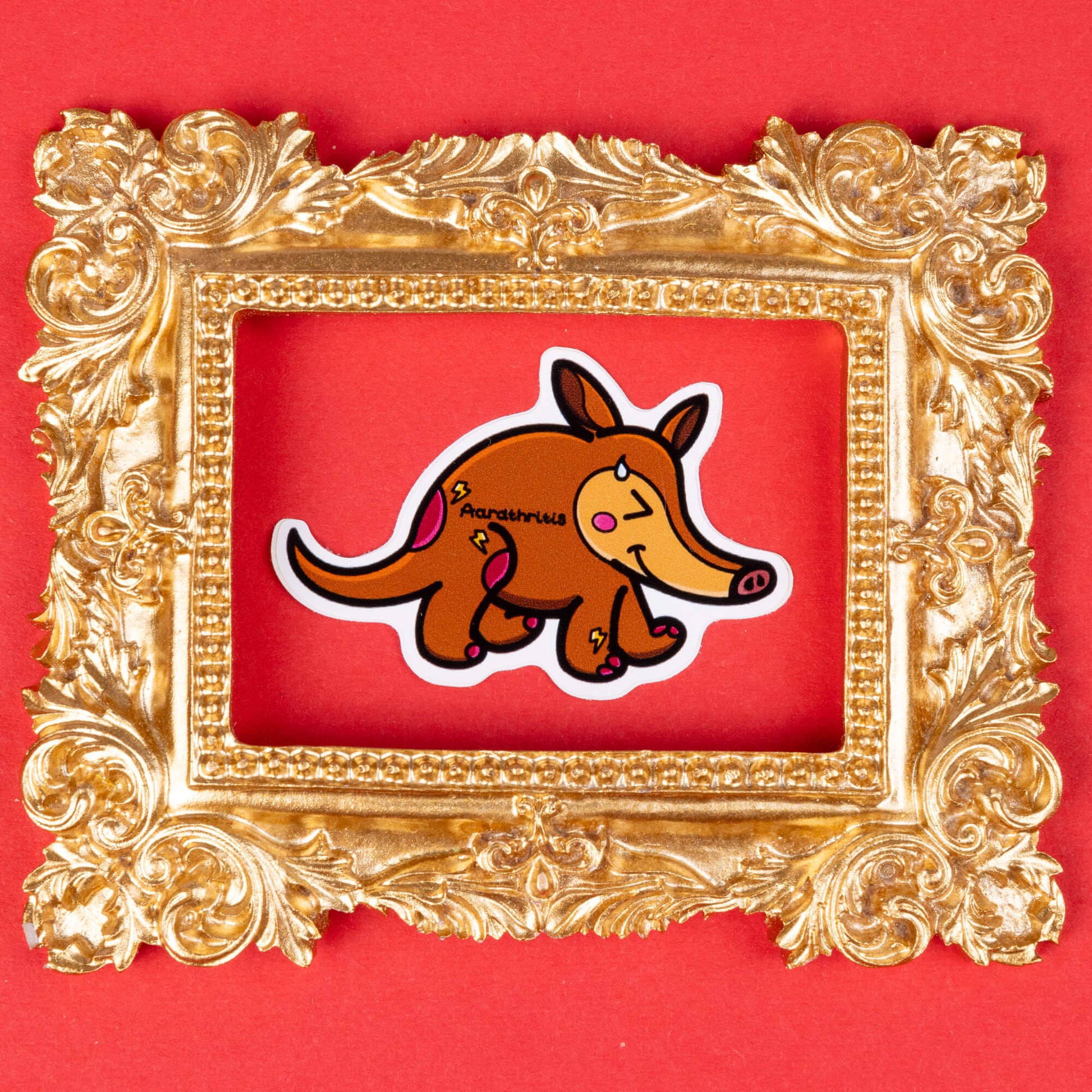 A cartoon sticker of a brown aardvark with a cheerful expression, blushing cheeks, and a drop of sweat on its face. The aardvark has pink spots and lightning bolt shapes on its body, with the word "Aardthritis" written on its side. The sticker is framed by an ornate, gold decorative frame and placed on a solid red background. Hand drawn design to raise awareness for arthritis.