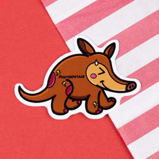 A cartoon sticker of a brown aardvark with a cheerful expression, blushing cheeks, and a drop of sweat on its face. The aardvark has pink spots and lightning bolt shapes on its body, with the word "Aardthritis" written on its side. The sticker is placed on a red background with a diagonal striped pink and white paper bag in the corner. Hand drawn design to raise awareness for arthritis.