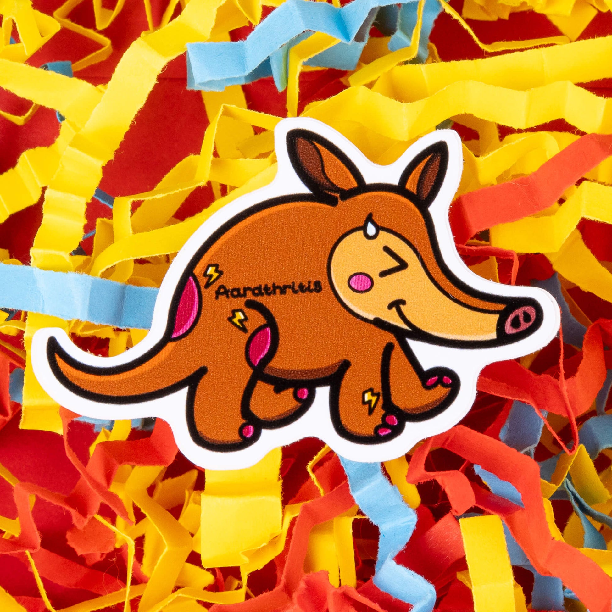 A cartoon sticker of a brown aardvark with a cheerful expression, blushing cheeks, and a drop of sweat on its face. The aardvark has pink spots and lightning bolt shapes on its body, with the word "Aardthritis" written on its side. The sticker is placed on a background of colorful shredded paper in yellow, red, and blue. Hand drawn design to raise awareness for arthritis.