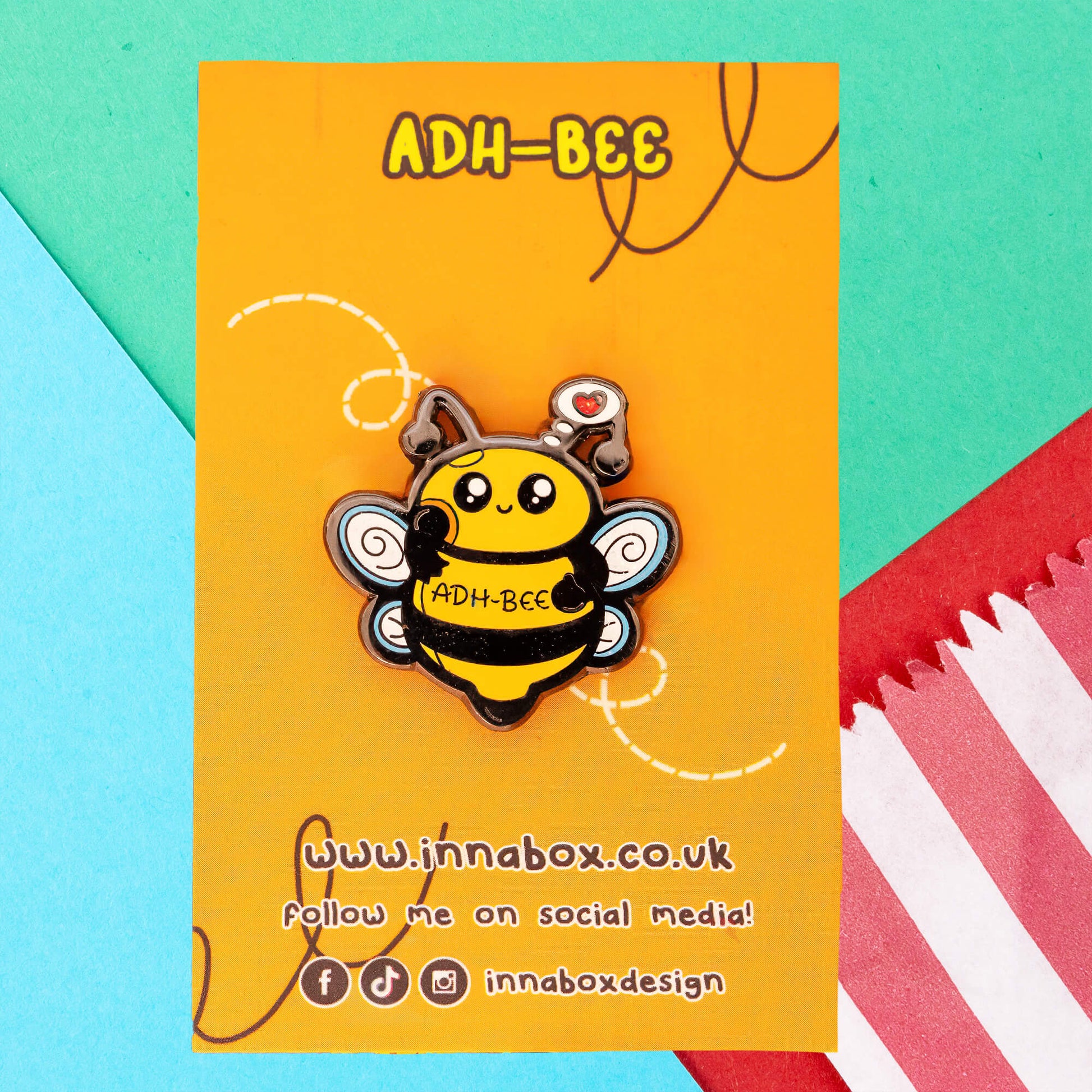 Innabox's ADH-Bee Glittery Bee Enamel Pin displayed on a bright orange backing card with a playful bee trail design. The pin features a smiling yellow and black bee with the text "ADH-Bee" on its body, cute swirly wings, and an antenna with a heart symbol.