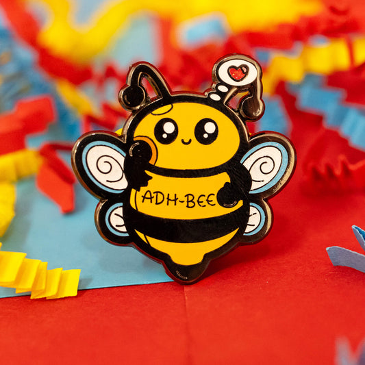 Close-up of the ADH-Bee Glittery Bee Enamel Pin by Innabox. This cheerful yellow and black bee has swirly wings, a heart-topped antenna, and "ADH-Bee" written across its body. Set against red, blue, and yellow crepe paper for a vibrant, playful background.