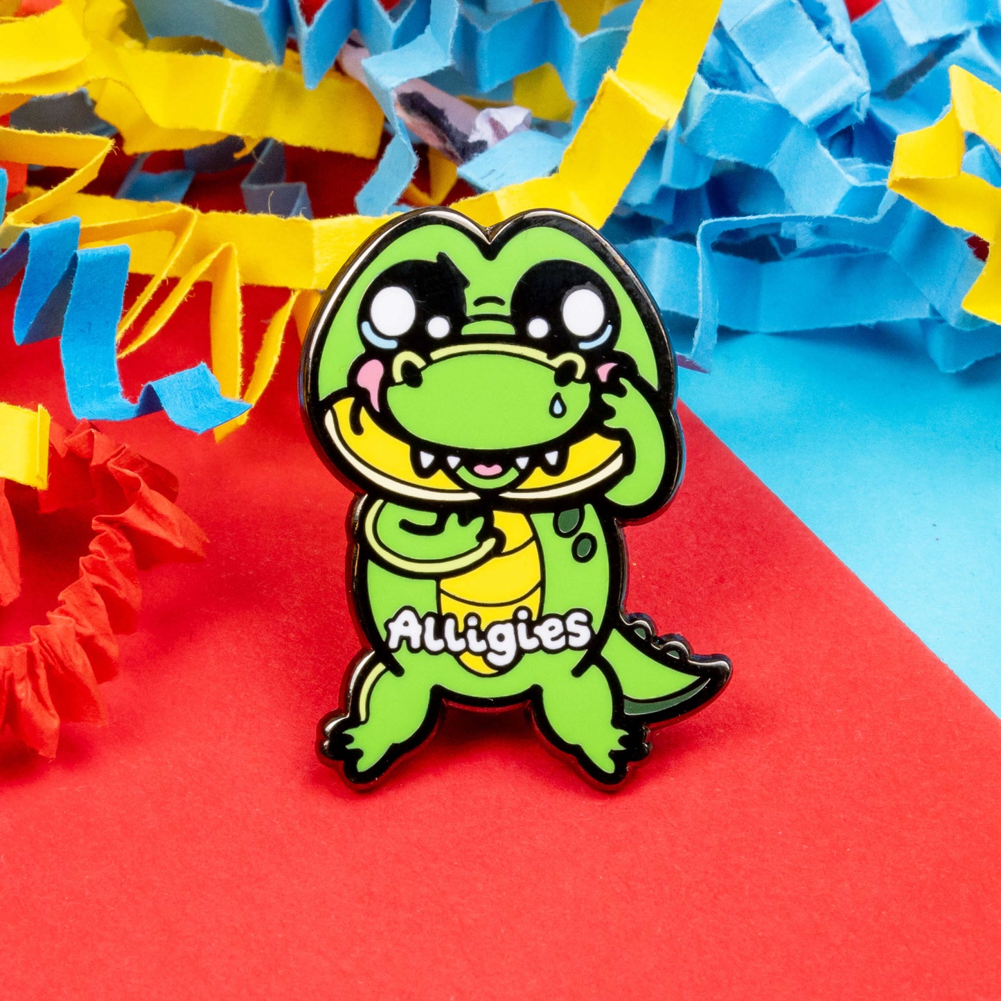 The Alligies Alligator Enamel Pin - Allergies on a red, blue and yellow card background. The green alligator shape pin has a sniffly nose, red puffy cheeks, holding up its front legs with white text across the front reading 'alligies'. The hand drawn design is raising awareness for allergies and allergens such as hay fever or anaphylaxis.