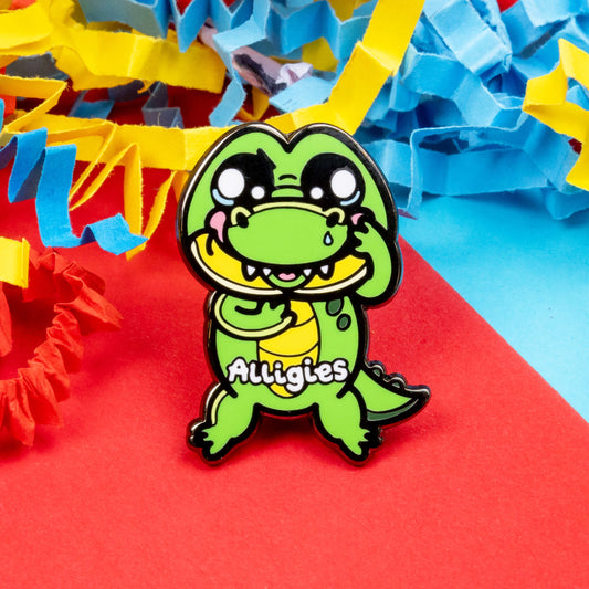 The Alligies Alligator Enamel Pin - Allergies on a red, blue and yellow card background. The green alligator shape pin has a sniffly nose, red puffy cheeks, holding up its front legs with white text across the front reading 'alligies'. The hand drawn design is raising awareness for allergies and allergens such as hay fever or anaphylaxis.