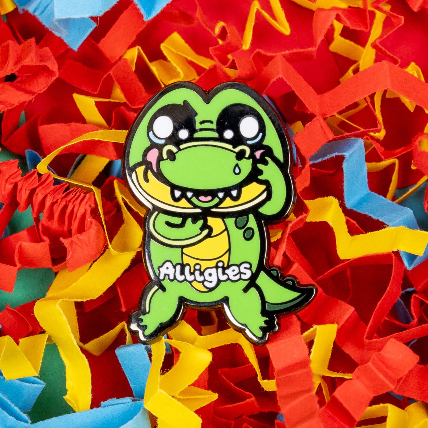 The Alligies Alligator Enamel Pin - Allergies on a red, blue and yellow card background. The green alligator shape pin has a sniffly nose, red puffy cheeks, holding up its front legs with white text across the front reading 'alligies'. The hand drawn design is raising awareness for allergies and allergens such as hay fever or anaphylaxis.