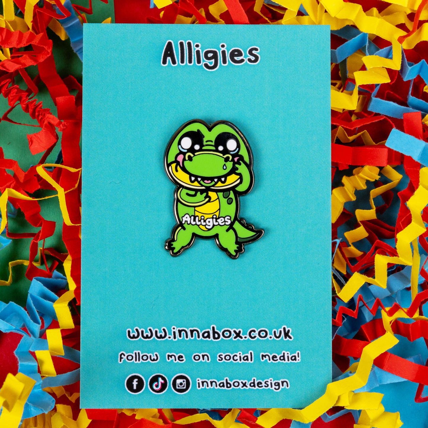 The Alligies Alligator Enamel Pin - Allergies on blue backing card sat on a red, blue and yellow card background. The green alligator shape pin has a sniffly nose, red puffy cheeks, holding up its front legs with white text across the front reading 'alligies'. The hand drawn design is raising awareness for allergies and allergens such as hay fever or anaphylaxis.