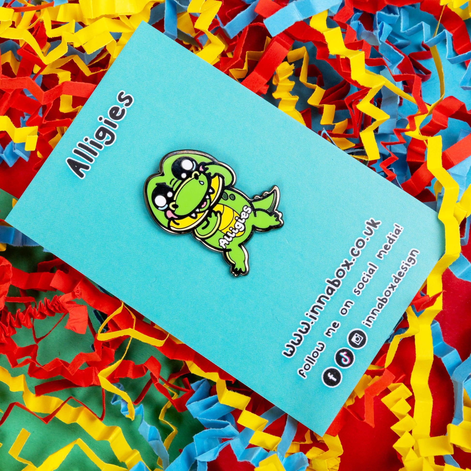 The Alligies Alligator Enamel Pin - Allergies on blue backing card sat on a red, blue and yellow card background. The green alligator shape pin has a sniffly nose, red puffy cheeks, holding up its front legs with white text across the front reading 'alligies'. The hand drawn design is raising awareness for allergies and allergens such as hay fever or anaphylaxis.