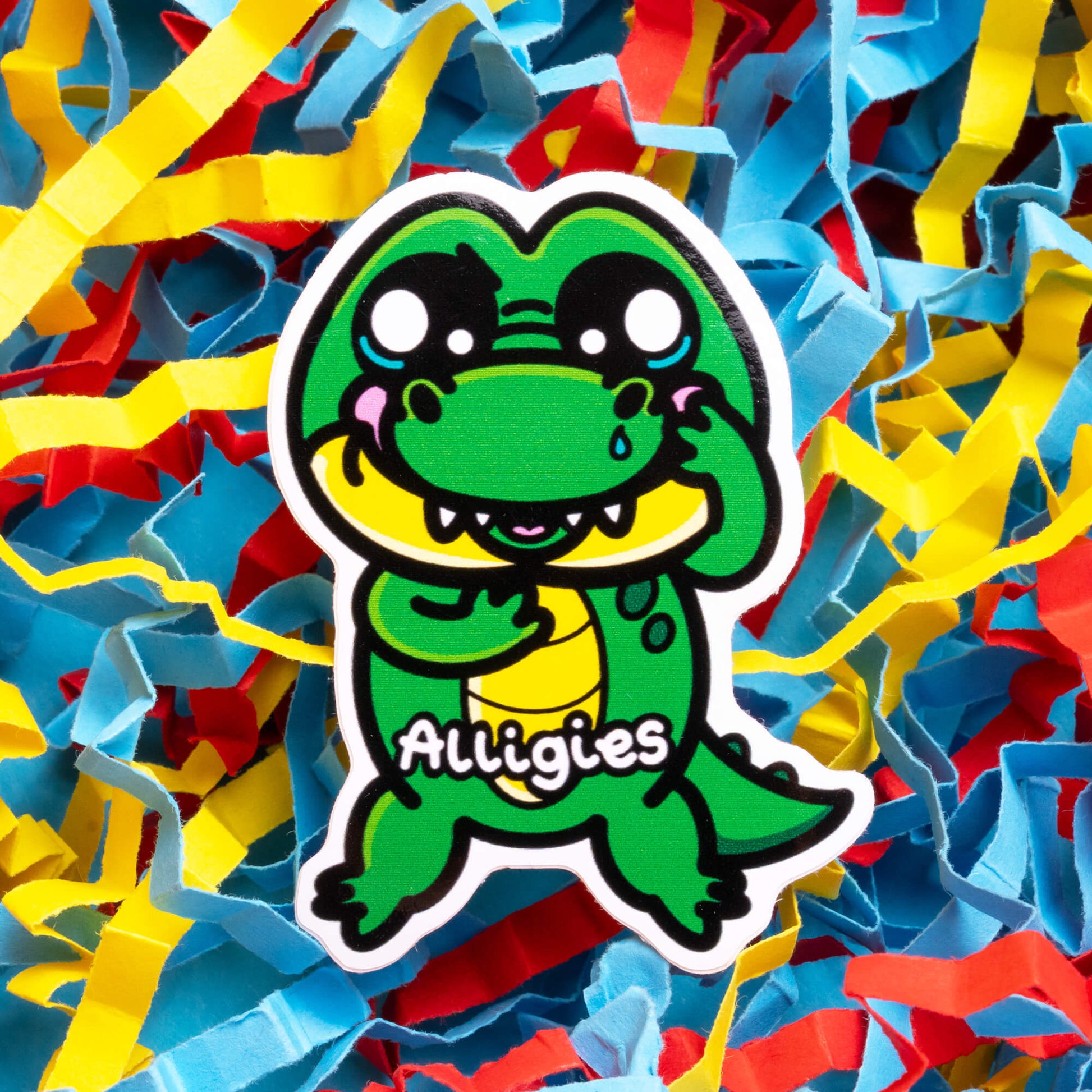 Innabox sticker featuring a cheerful green alligator character named 'Alligies,' with large, expressive eyes and a yellow belly. The character represents allergies, designed to bring comfort to those with chronic illnesses or hidden disabilities. The alligator stands out against a colourful background of blue, red, and yellow paper streamers, symbolising celebration and resilience.