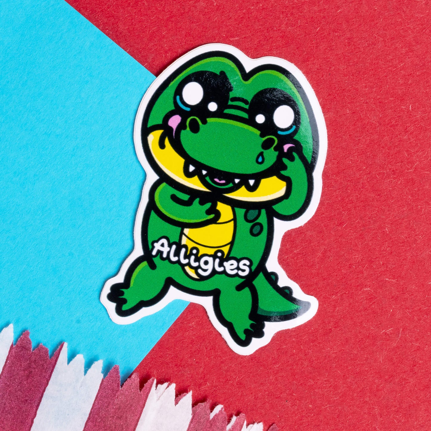 Innabox's playful sticker design featuring a cute green alligator with big, teary eyes, holding its cheeks. The text 'Alligies' is displayed on the character's belly, representing allergies in a fun and uplifting way. The background consists of a vibrant mix of red, blue, and white colours, adding a dynamic feel to the design. Perfect for individuals with chronic illnesses or hidden disabilities.