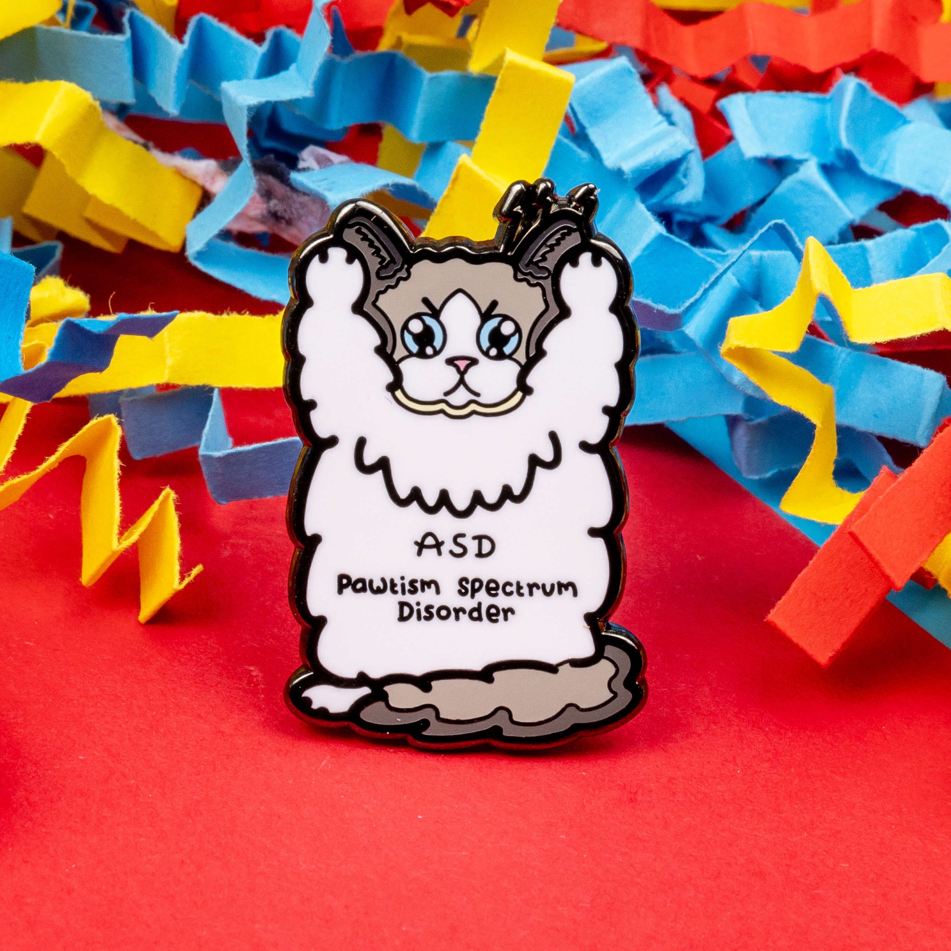 Pawtism Spectrum Disorder Cat Enamel Pin - Autism Spectrum Disorder (ASD) on a red background with blue, yellow and red card confetti around it. The pin is of a grey and white cat with a stern expression and paws in the air. 'ASD Pawtism Spectrum Disorder' is written across it's stomach in black text. Hand drawn design to raise awareness for Autism Spectrum Disorder (ASD)