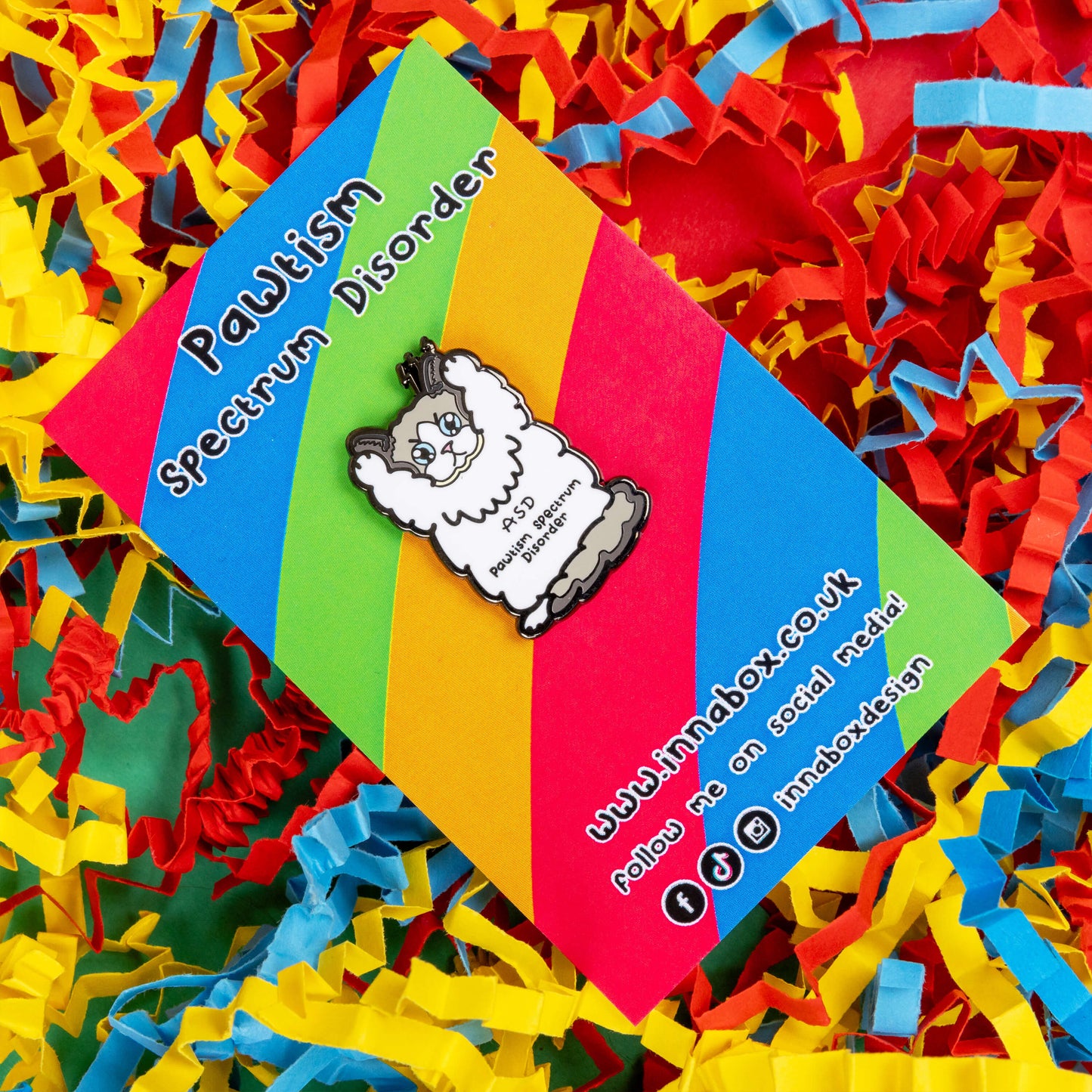 Pawtism Spectrum Disorder Cat Enamel Pin - Autism Spectrum Disorder (ASD) on it's rainbow striped backing card with Innabox social media handles written on it. It is resting on a blue, yellow and red coloured card confetti background. The pin is of a grey and white cat with a stern expression and paws in the air. 'ASD Pawtism Spectrum Disorder' is written across it's stomach in black text. Hand drawn design to raise awareness for Autism Spectrum Disorder (ASD)