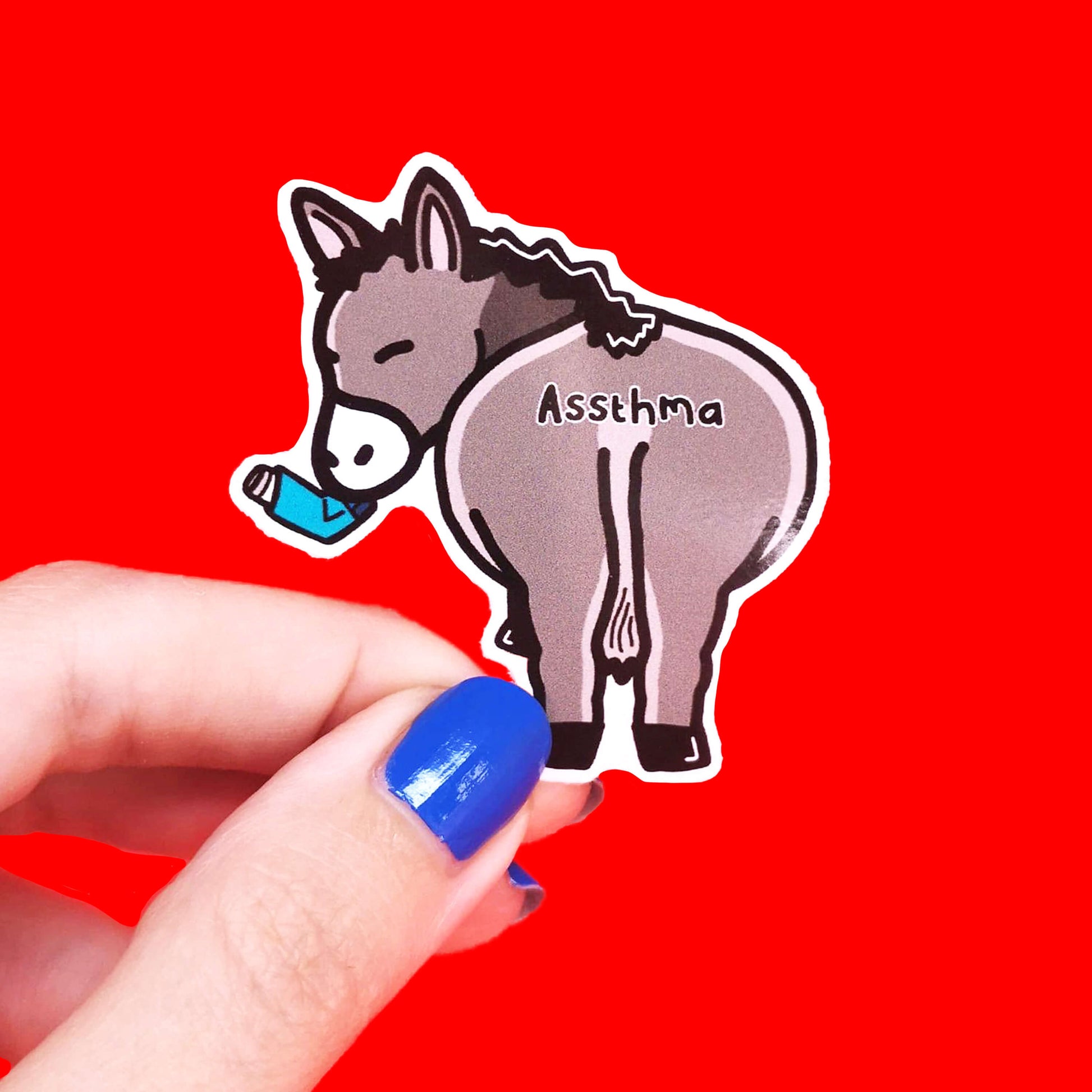 Assthma Sticker - Asthma being held over a red background by a hand with blue nail varnish. A grey donkey ass showing its behind with a blue asthma pump in its mouth and the word Assthma across its behind. The sticker is designed to raise awareness for asthma.