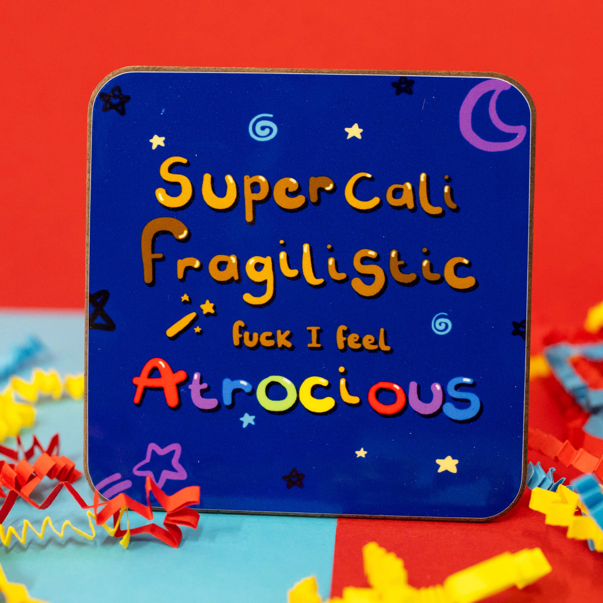 Close-up of the I Feel Atrocious Coaster by Innabox. The square coaster has rounded corners and a deep blue background decorated with stars, swirls, and a purple crescent moon. The text reads, "SuperCaliFragilistic f*** I feel Atrocious" in playful, multicoloured letters. Surrounding the coaster are red, yellow, and blue crepe paper decorations.