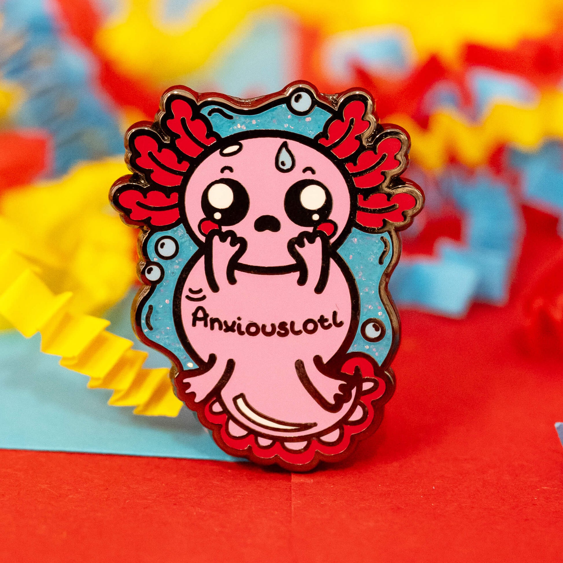 Close-up of the Anxiouslotl Glittery Axolotl Enamel Pin by Innabox. The pin features a pink axolotl with red gills, big anxious eyes, and flushed cheeks, holding its face in worry. It rests on red, blue, and yellow crinkle paper, with a glittery blue background surrounding the character.