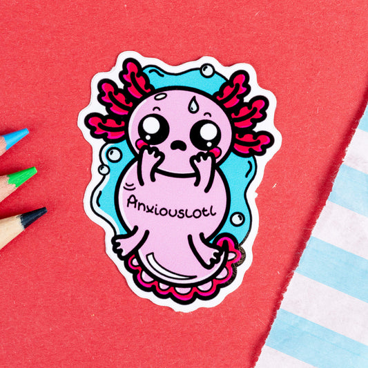 Close-up of the Anxiouslotl Axolotl Sticker by Innabox. The sticker features a worried pink axolotl with big, teary eyes, flushed cheeks, and red gills. It’s holding its face with tiny hands, and the word "Anxiouslotl" is written across its body. The background is a bubbly blue outline, and it's set against a red surface with coloured pencils nearby.