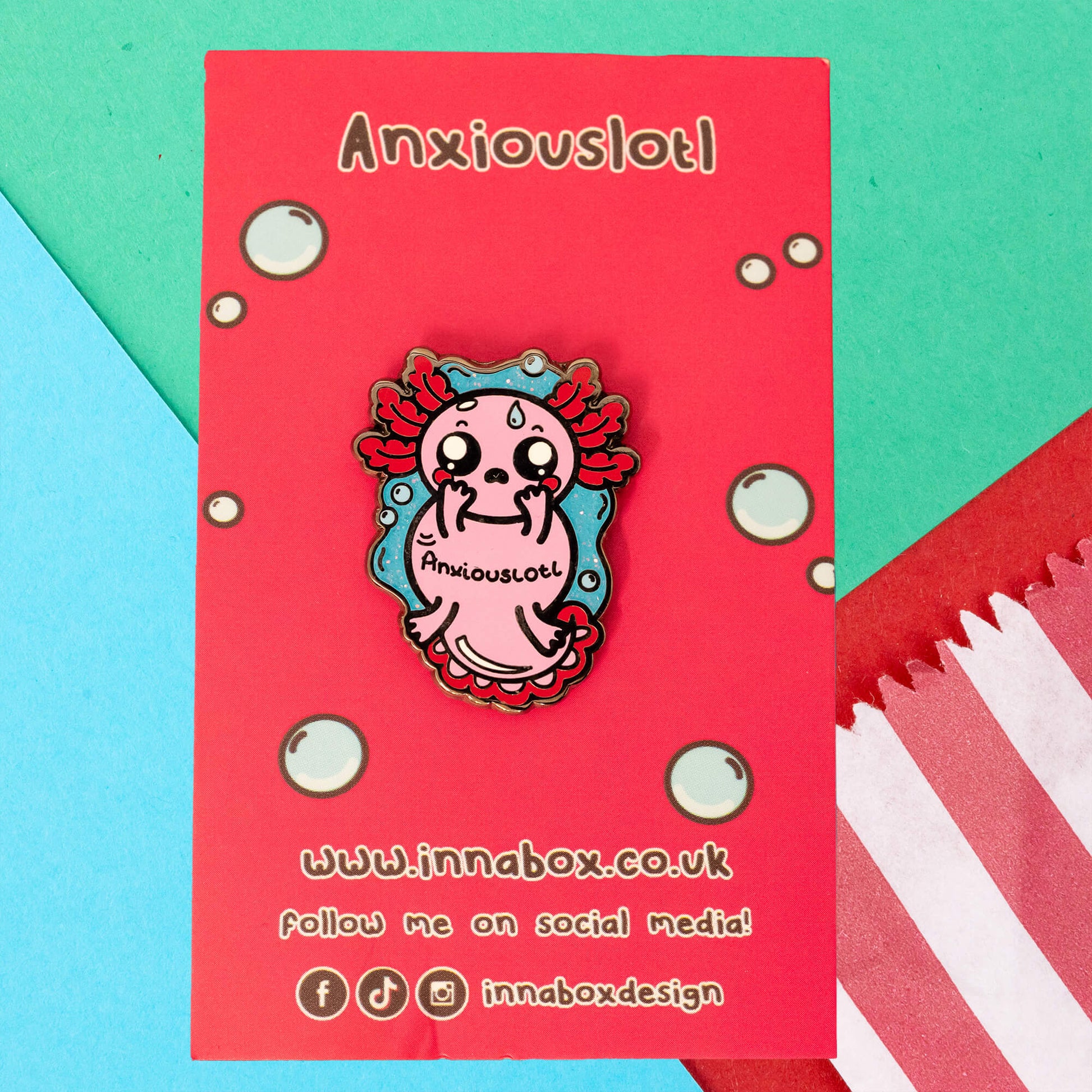 Innabox's Anxiouslotl Glittery Axolotl Enamel Pin displayed on a bright red backing card with bubble illustrations and the word "Anxiouslotl" at the top. The pin features a pink axolotl with wide, worried eyes, flushed cheeks, and red gills, holding its face with tiny hands.