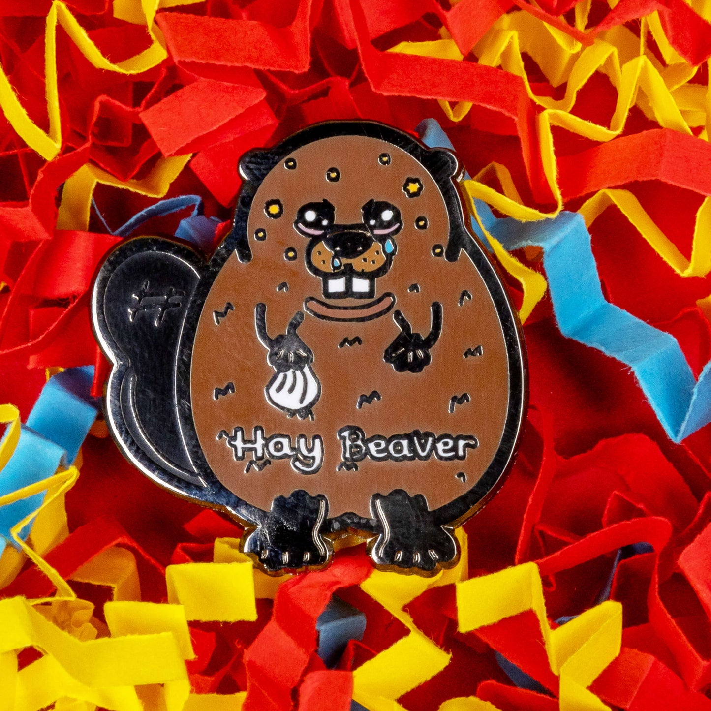 Hay Beaver Enamel Pin - Hay Fever on a blue, yellow and red coloured card confetti background. The enamel pin is a brown beaver with watery eyes, dripping nose and yellow spots on face and is holding a tissue with it's little hand. Hay beaver is written across its belly. The enamel pin is designed to raise awareness for hay fever or allergic rhinitis
