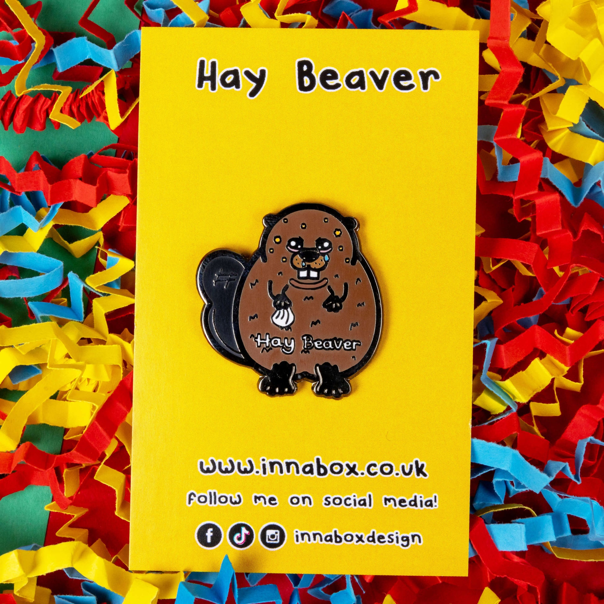 Hay Beaver Enamel Pin - Hay Fever on yellow backing in front of blue, yellow and red coloured card confetti background. The enamel pin is a brown beaver with watery eyes, dripping nose and yellow spots on face and is holding a tissue with it's little hand. Hay beaver is written across its belly. The enamel pin is designed to raise awareness for hay fever or allergic rhinitis