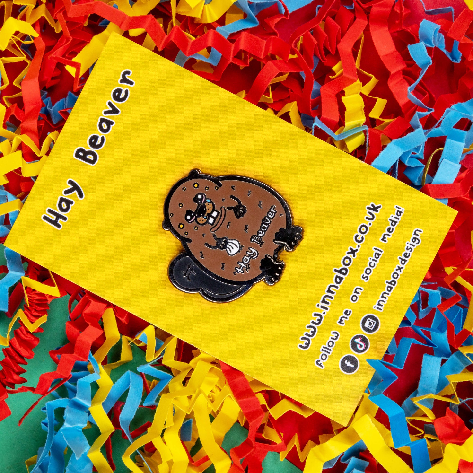 Hay Beaver Enamel Pin - Hay Fever on yellow backing in front of blue, yellow and red coloured card confetti background. The enamel pin is a brown beaver with watery eyes, dripping nose and yellow spots on face and is holding a tissue with it's little hand. Hay beaver is written across its belly. The enamel pin is designed to raise awareness for hay fever or allergic rhinitis
