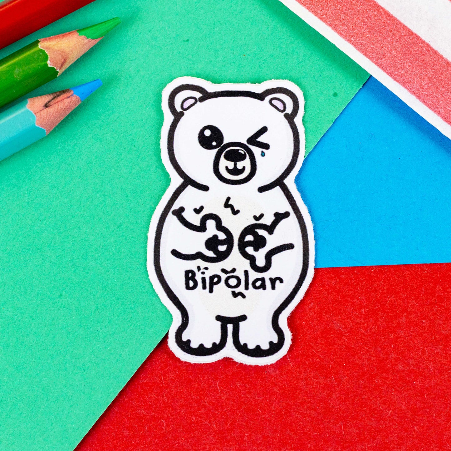 Bipolar Bear Sticker - Bipolar on a red, blue and green background with colouring pencils and a red stripe candy bag. The sticker is a white polar bear with one eye closed crying whilst smiling clutching its chest, across its tummy reads bipolar. The pun sticker is raising awareness for bipolar disorder.
