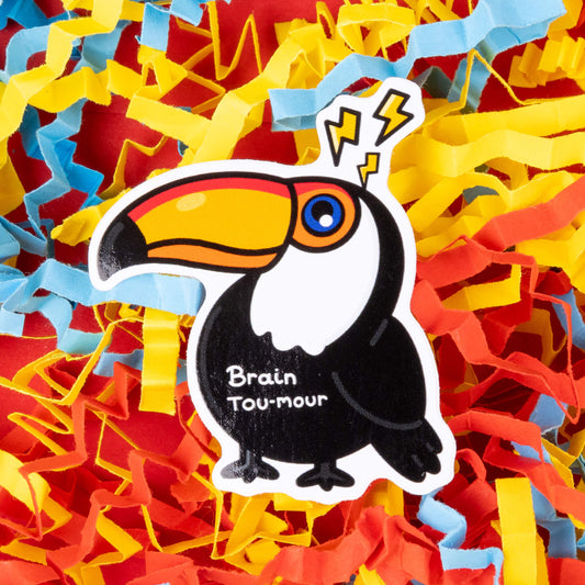 A sticker of a toucan with a vibrant orange beak and a black and white body. The toucan has lightning bolts above its head and the text "Brain Tou-mour" written on its chest. The sticker is placed on a background of colorful shredded paper in yellow, blue, and red. Hand drawn design created to raise awareness for brain tumours.