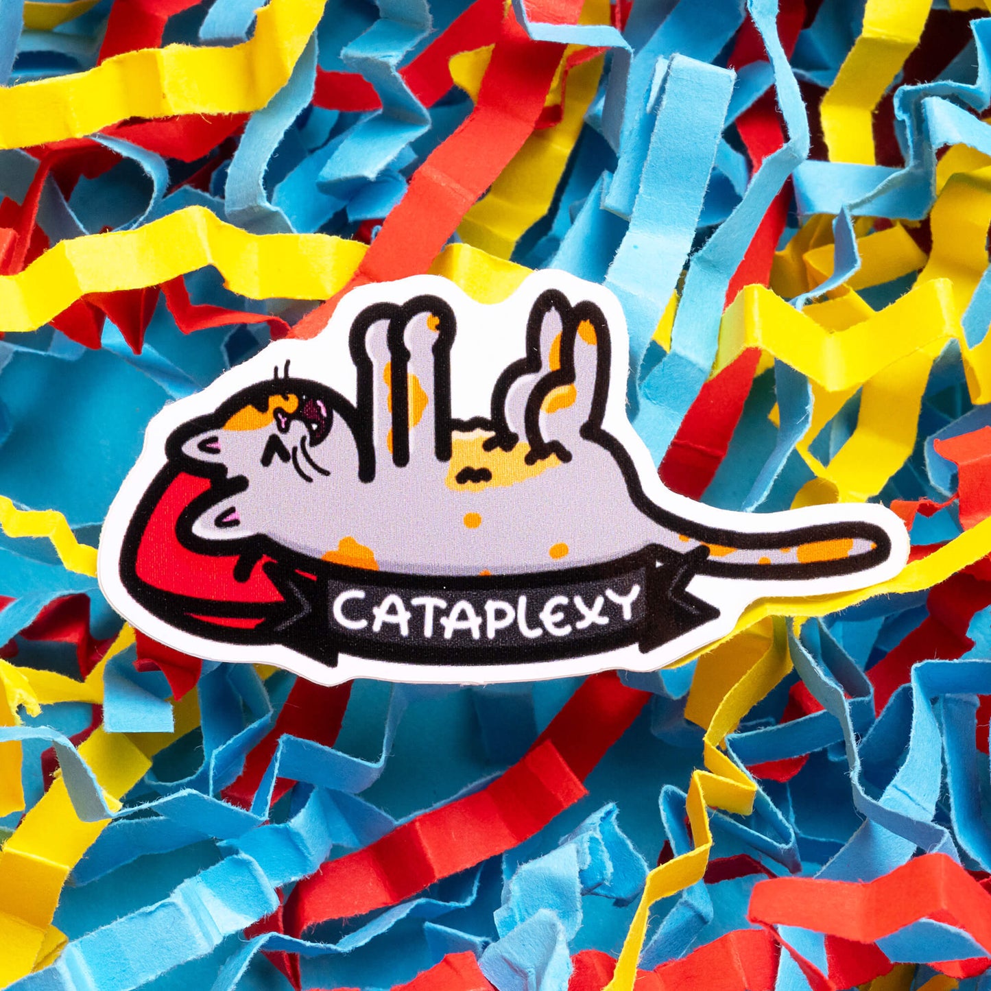 Colourful Innabox sticker featuring a cute cartoon cat in a playful, upside-down position with the text 'Cataplexy' below. The background is filled with vibrant, multicoloured paper streamers. Innabox is a brand known for creating disability-inspired products to uplift those with chronic illnesses and hidden disabilities.