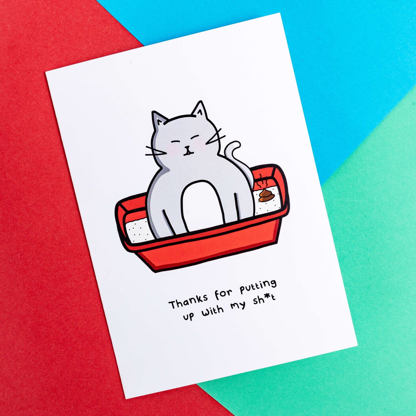 A white card with a drawing of a grey cat sat in a litter box with a poop in it with text underneath that says, thanks for putting up with my sh*t. The a6 thank you card is laying on a red, blue and green background.