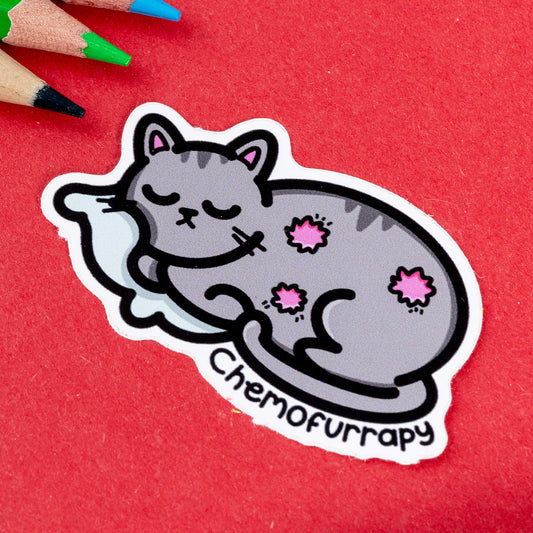 Close-up of the Chemofurrapy Cat Sticker by Innabox. The sticker features a grey cat with closed eyes, peacefully curled up on a white pillow. The cat has three pink, flower-like patches symbolising areas affected by chemotherapy. The word "Chemofurrapy" is written in bold, playful black text at the bottom. Set against a red background with coloured pencils nearby.